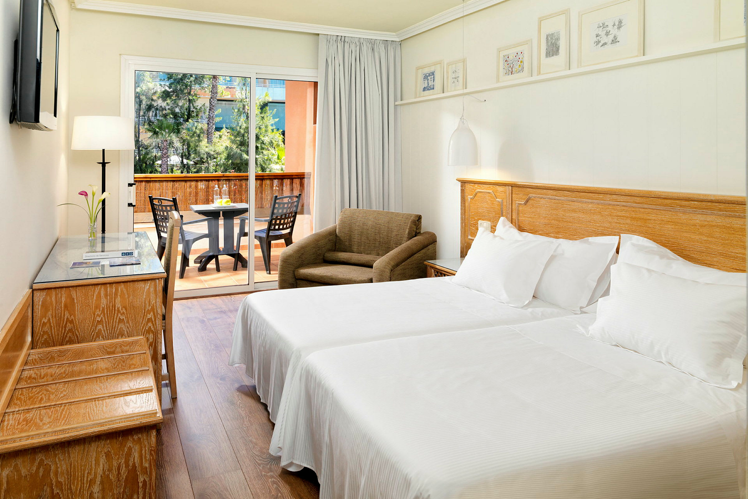 Ett hotellrum med två enkelsängar täckta av vita sängkläder. Det finns en fåtölj, ett skrivbord och en glasterrassdörr som leder till balkongen med ett bord och stolar. (AI-genererad text)