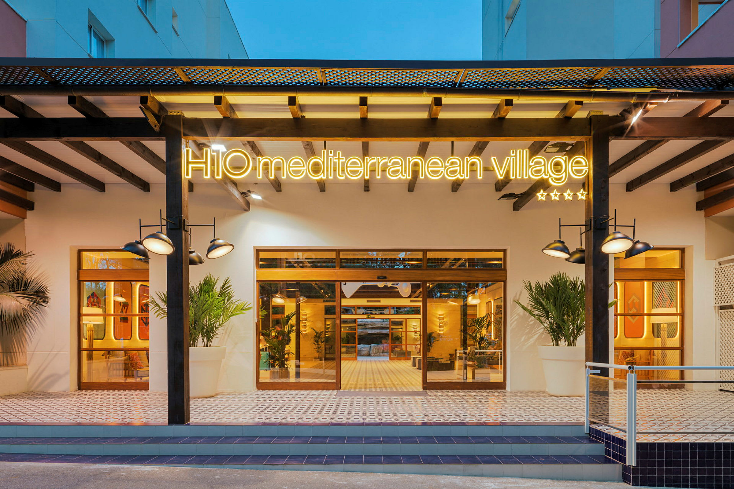 Entrén till H10 Mediterranean Village, ett hotell med belyst skylt, stora fönster och modern design. (AI-genererad text)