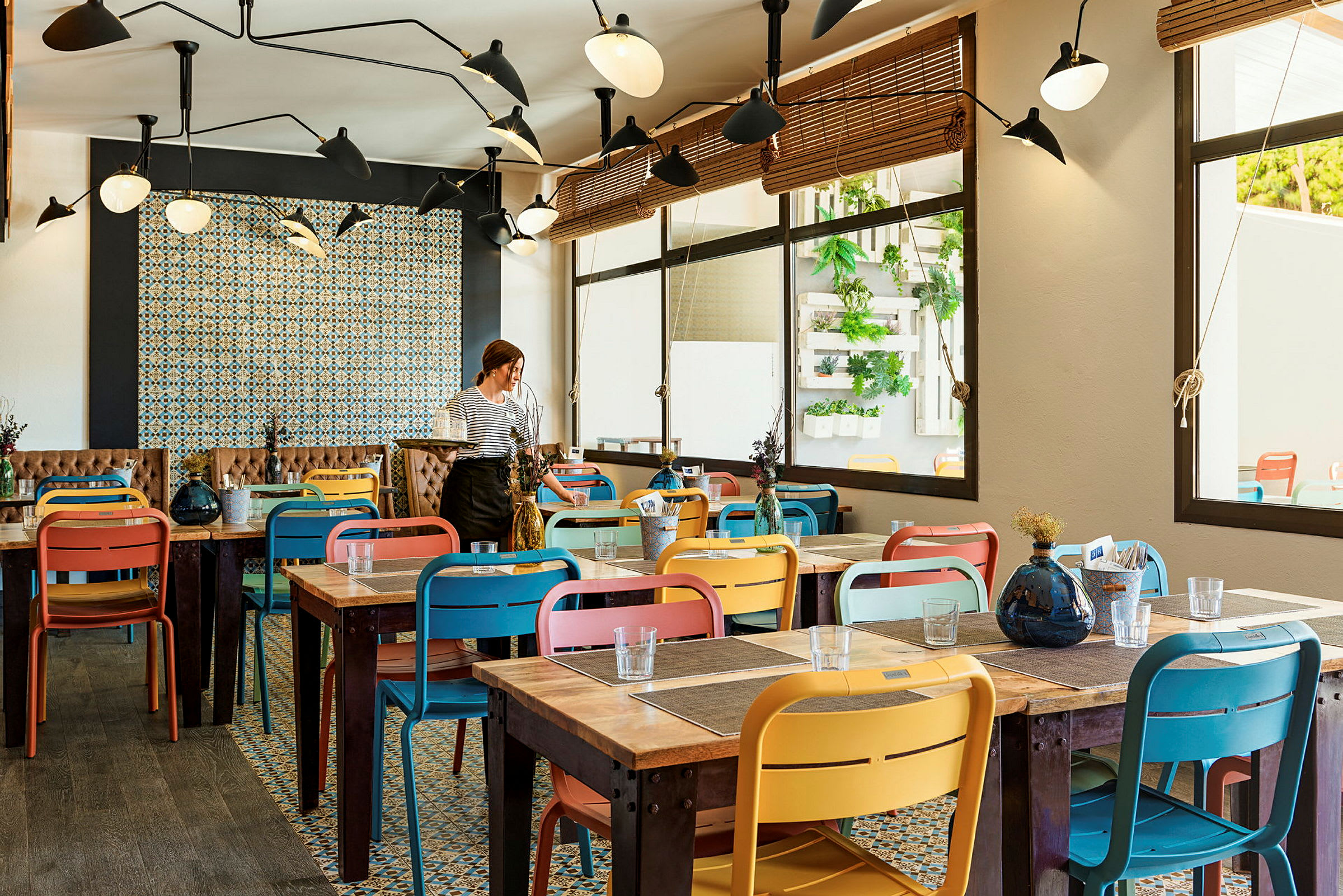 Inomhusbild från en restaurang med färgglada stolar och träbord. En person står vid ett av borden. (AI-genererad text)