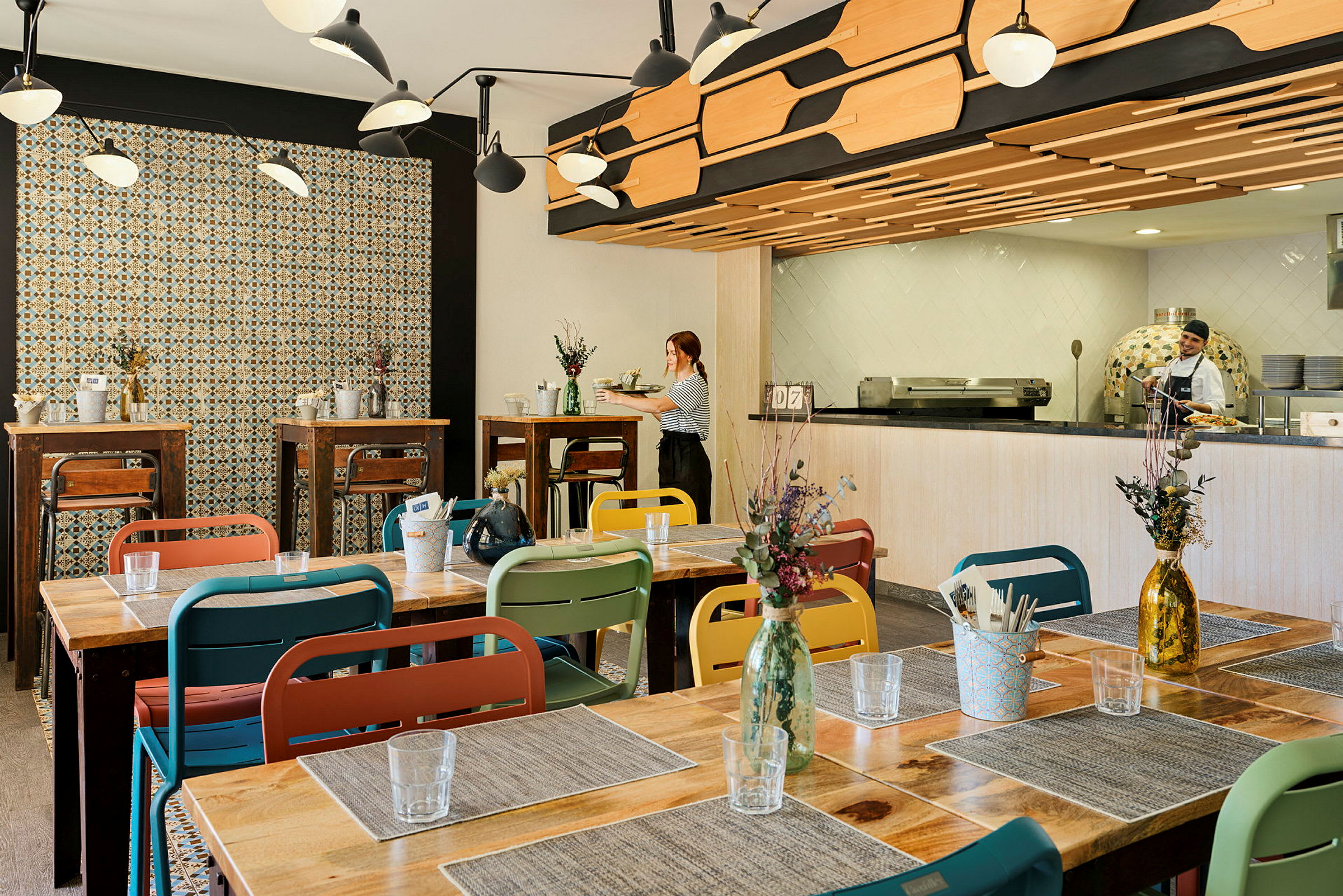 Bild av en restaurangmiljö med färgglada stolar och träbord, dekorerad med blommor i vaser. Väggarna har mönstrat kakel och köket är synligt i bakgrunden. (AI-genererad text)