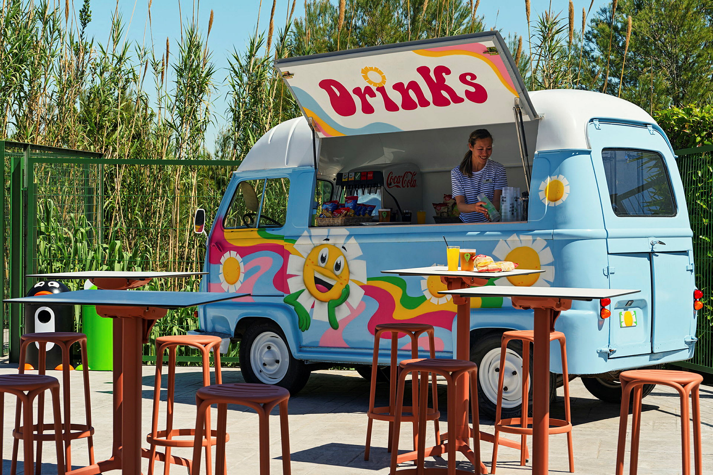 En färgglad matbil med texten 'Drinks' serverar drycker och snacks. Framför bilen står bord och stolar. (AI-genererad text)
