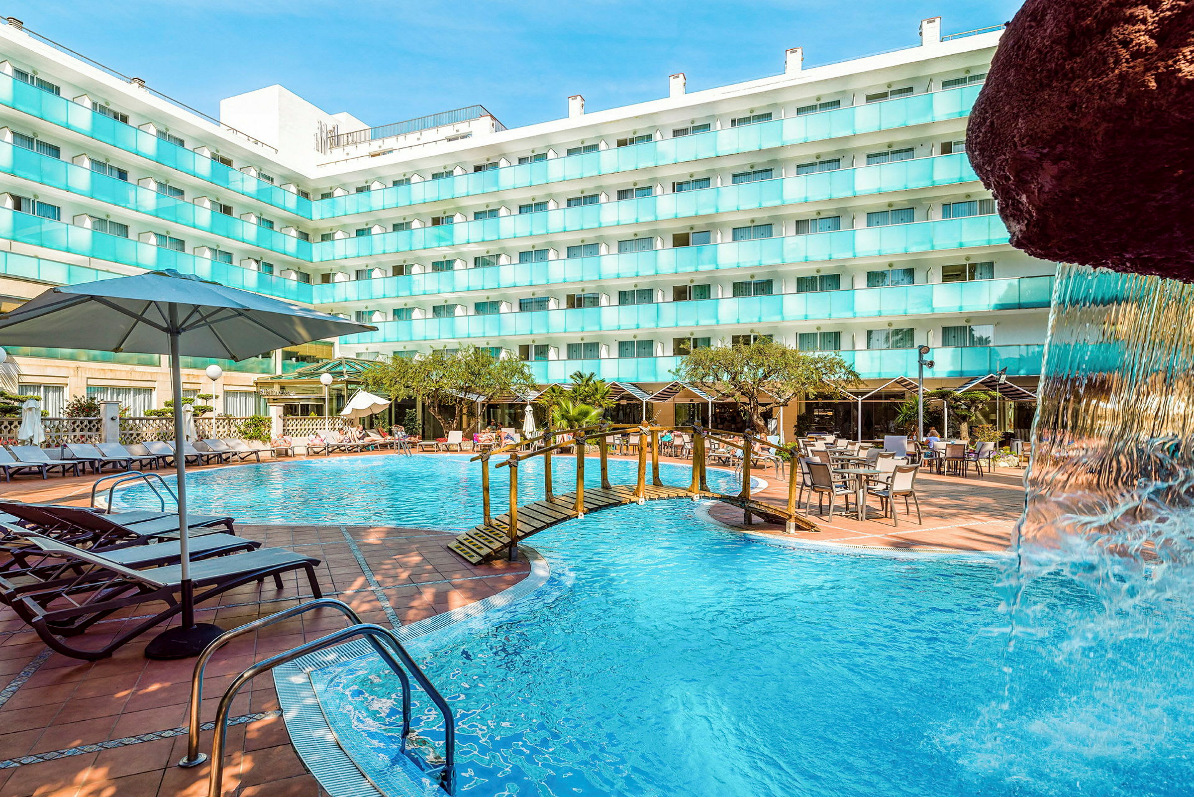 En utomhuspool vid en hotellbyggnad med solstolar, bord och parasoller. En liten bro över poolen och en del av en vattenfallstruktur syns. (AI-genererad text)