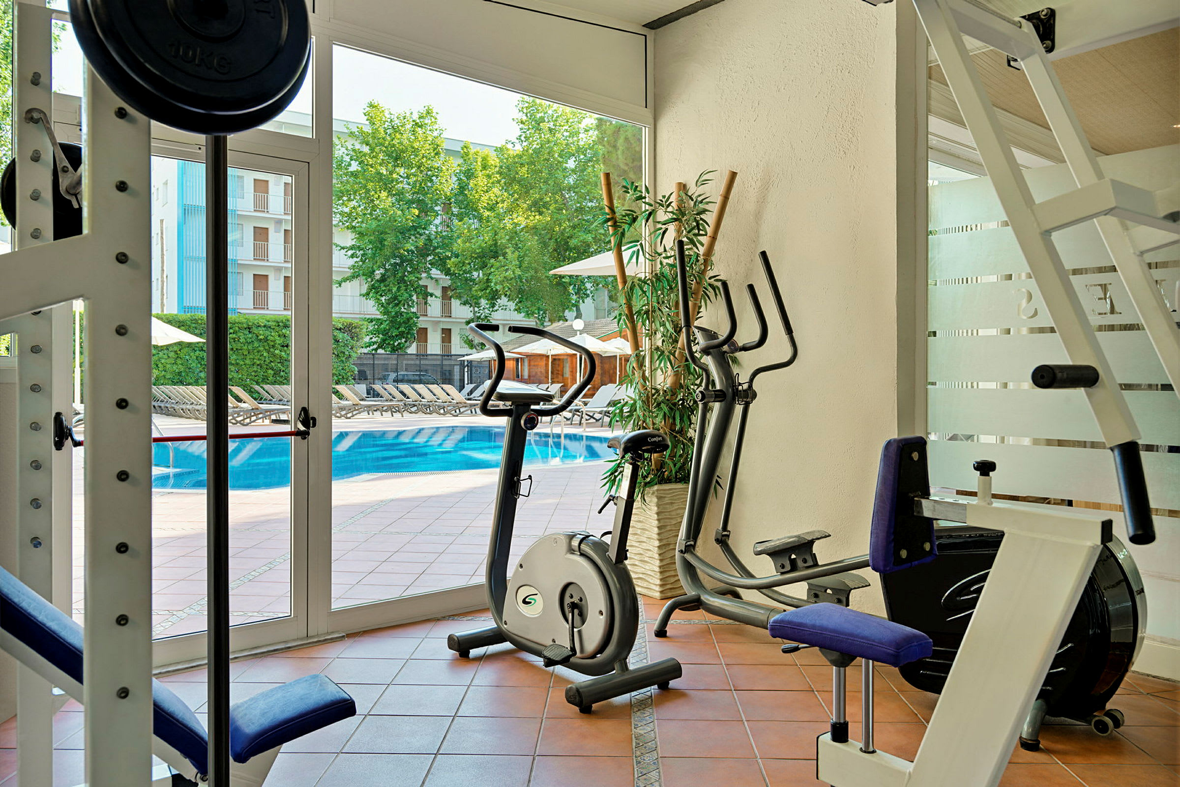 Ett gym med träningsmaskiner och motionscyklar, nära en pool utomhus. (AI-genererad text)
