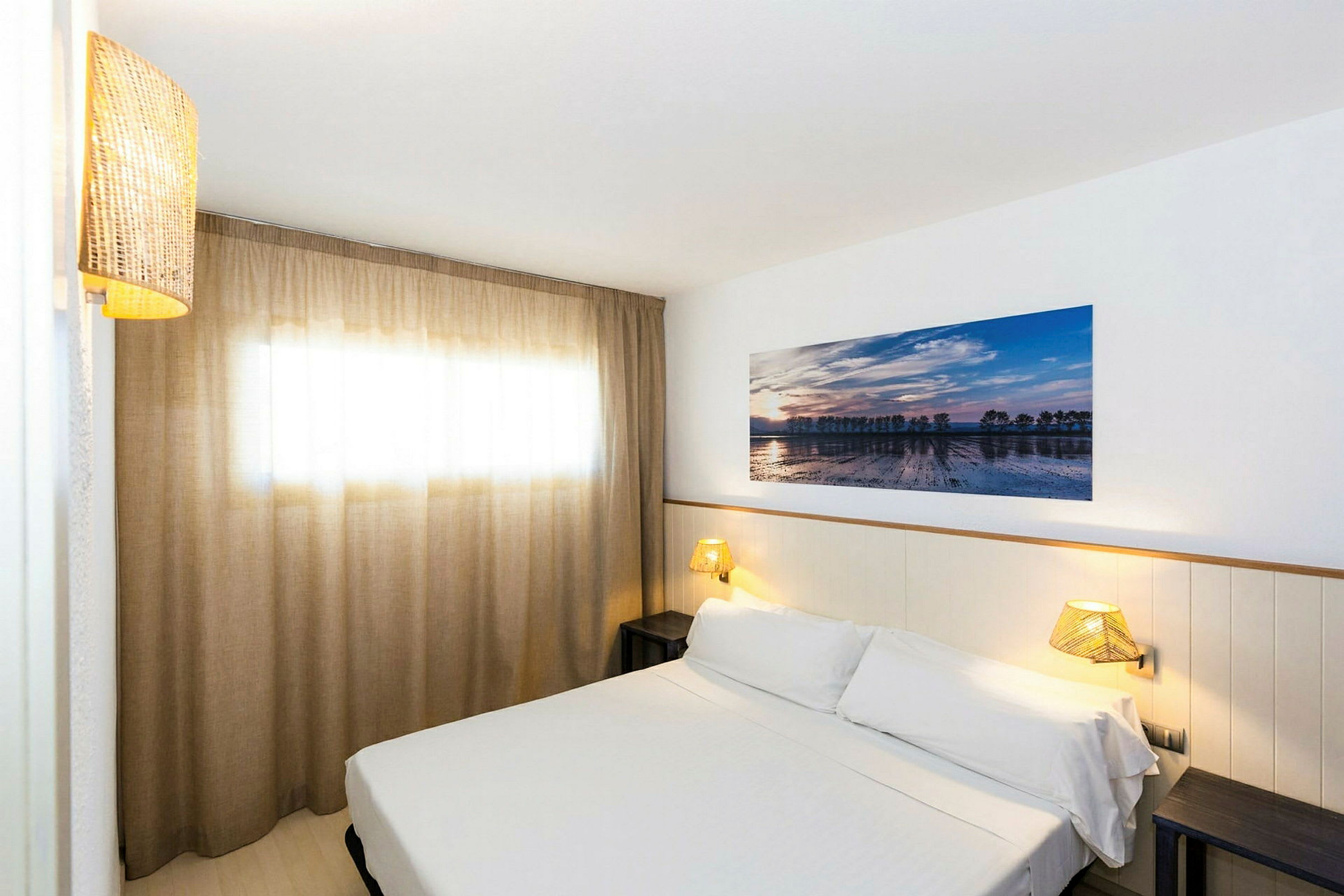 Ett hotellrum med en säng klädd i vitt linne, en vägglampa och ett konstverk på väggen. Ett fönster med eleganta beiga gardiner. (AI-genererad text)