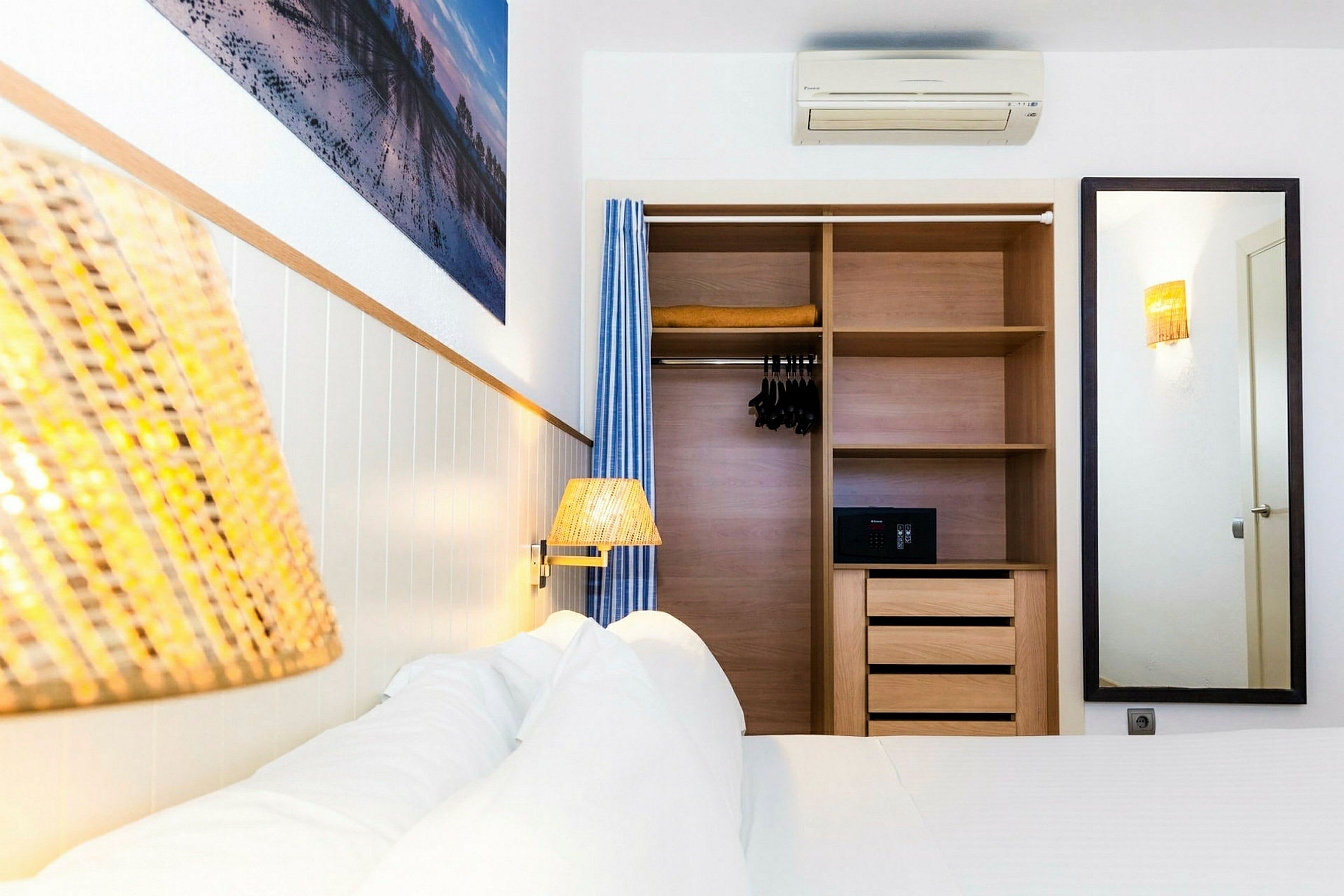 Ett hotellrum med en säng och en inbyggd garderob. Garderoben är öppen och innehåller hyllor, galgar och ett kassaskåp. På väggen syns en spegel och en tavla. (AI-genererad text)