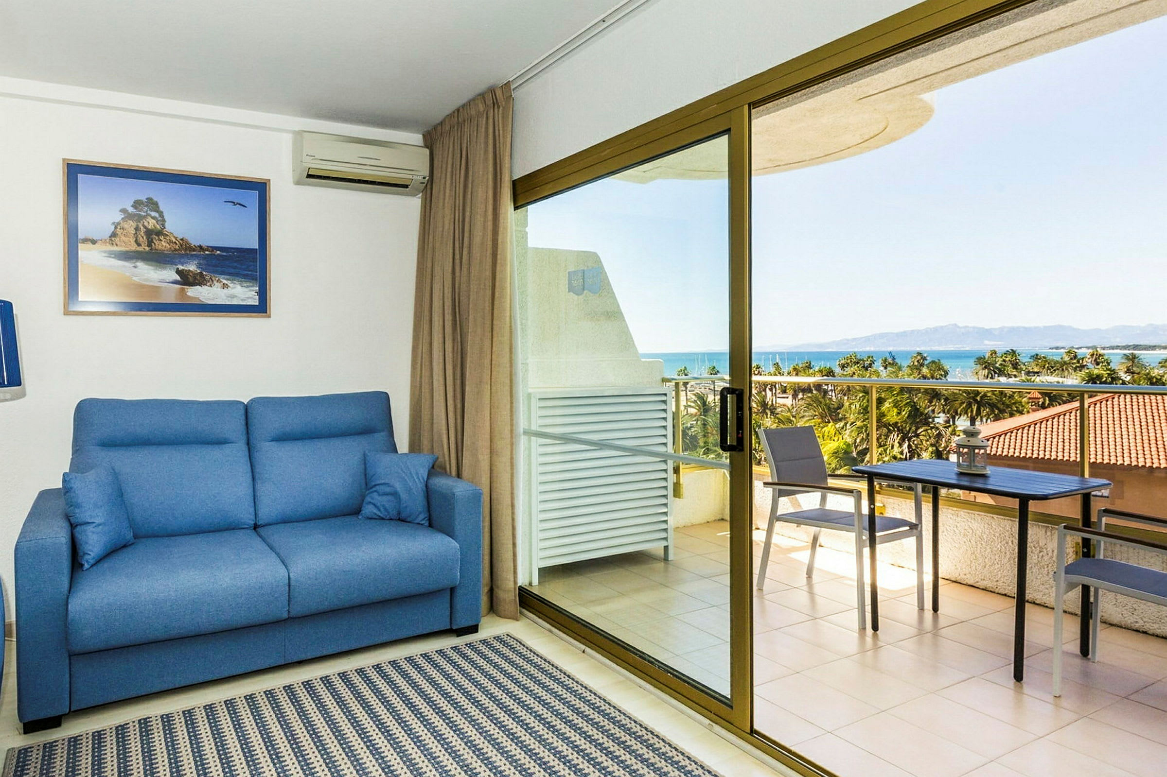 Ett rum med en blå soffa, matta och ett glastakfönster ut mot en terrass. Terrassen har bord, stolar och utsikt över havet. (AI-genererad text)