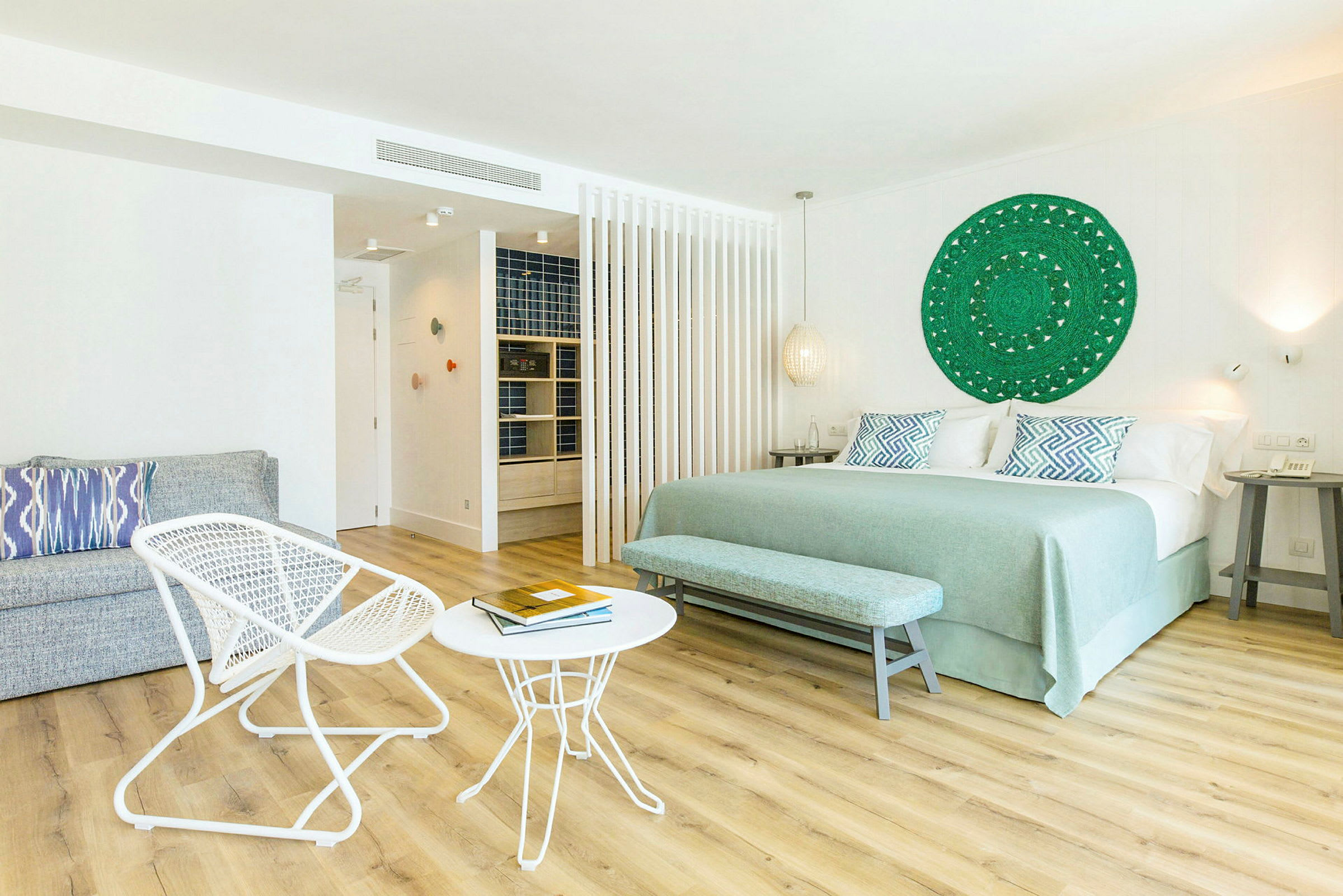 Ett ljust hotellrum med en stor säng, två små bord och två stolar, samt en dekorativ väggprydnad. (AI-genererad text)