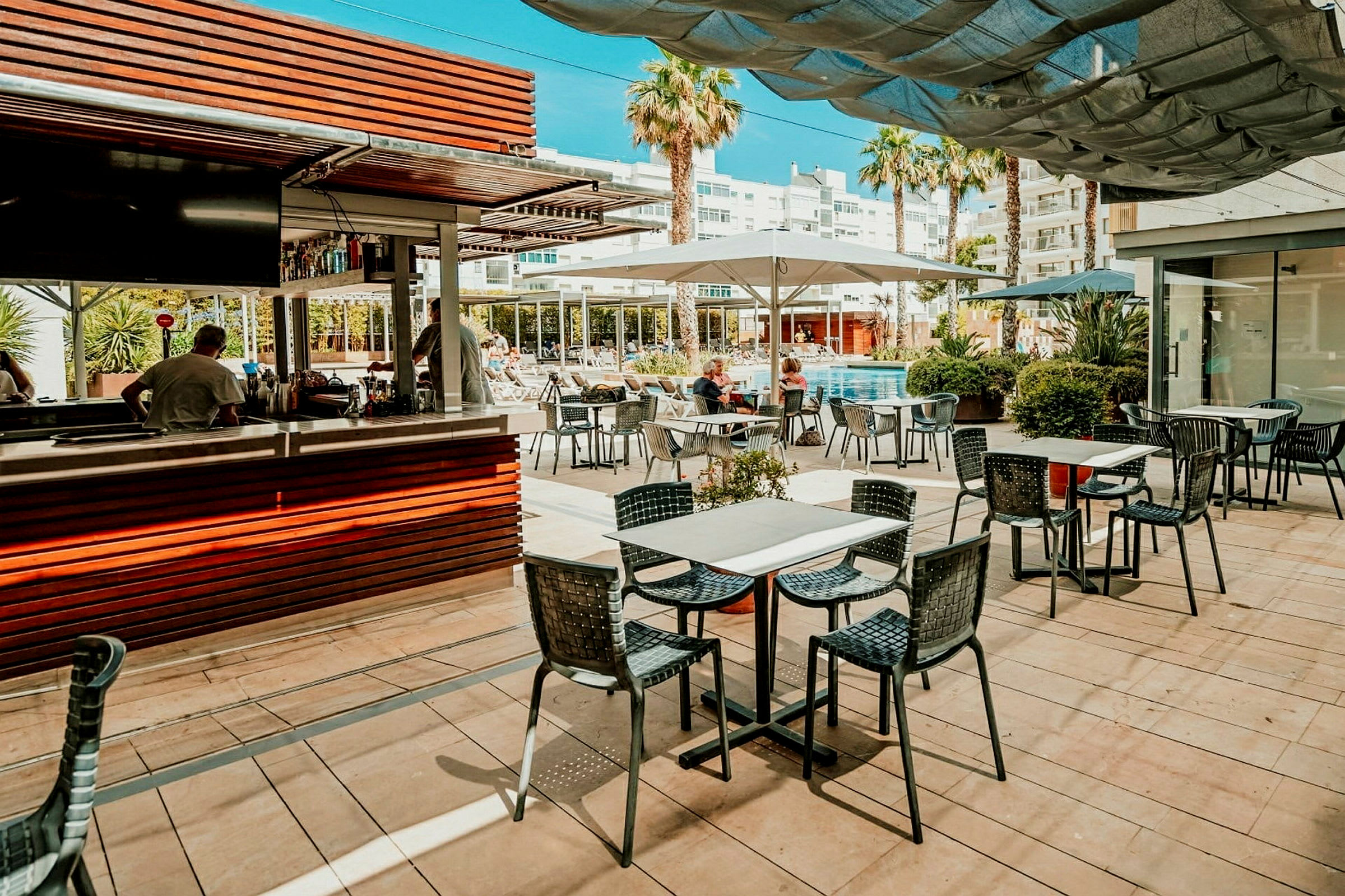 En poolnära restaurang med sittplatser utomhus och en serveringsdisk, inramad av omgivande byggnader och palmer. (AI-genererad text)