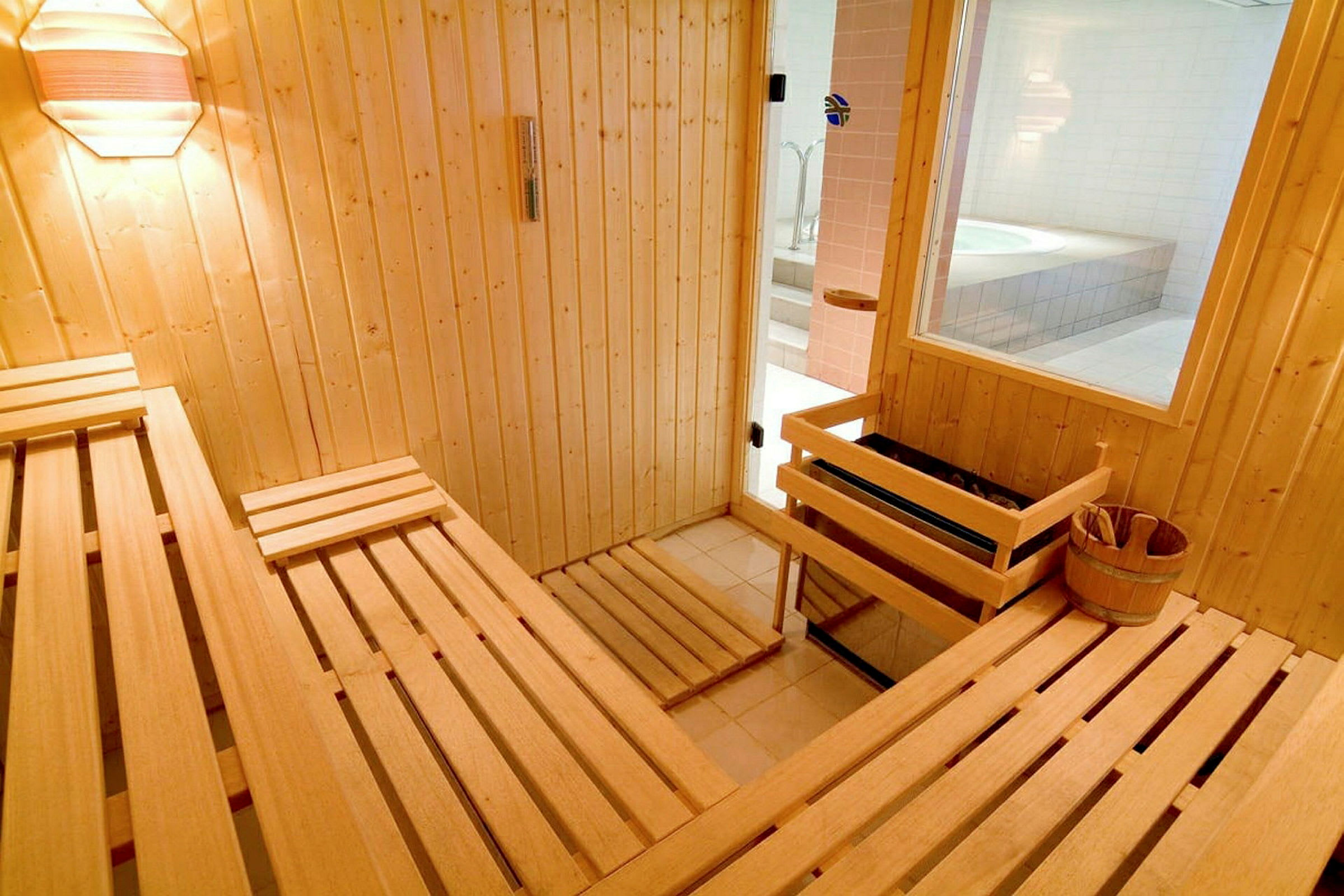 En träinredning i en bastu med bänkar. Ett fönster och en dörr vetter mot ett rum med ett bubbelbadkar. (AI-genererad text)