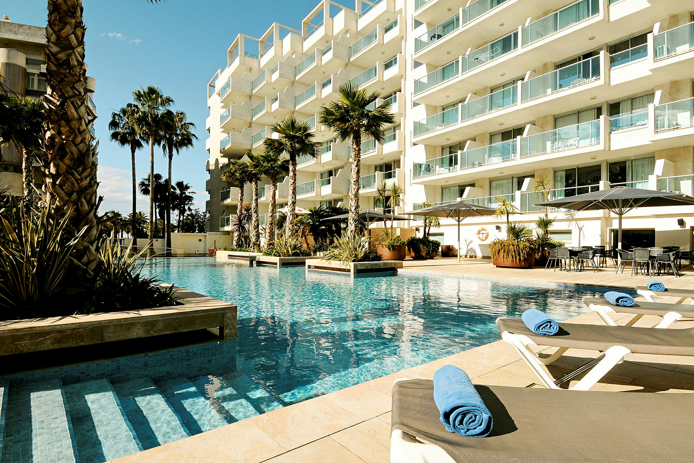 Bild av en hotellpool med solstolar och palmer omgivna av ett modern byggnadskomplex. (AI-genererad text)