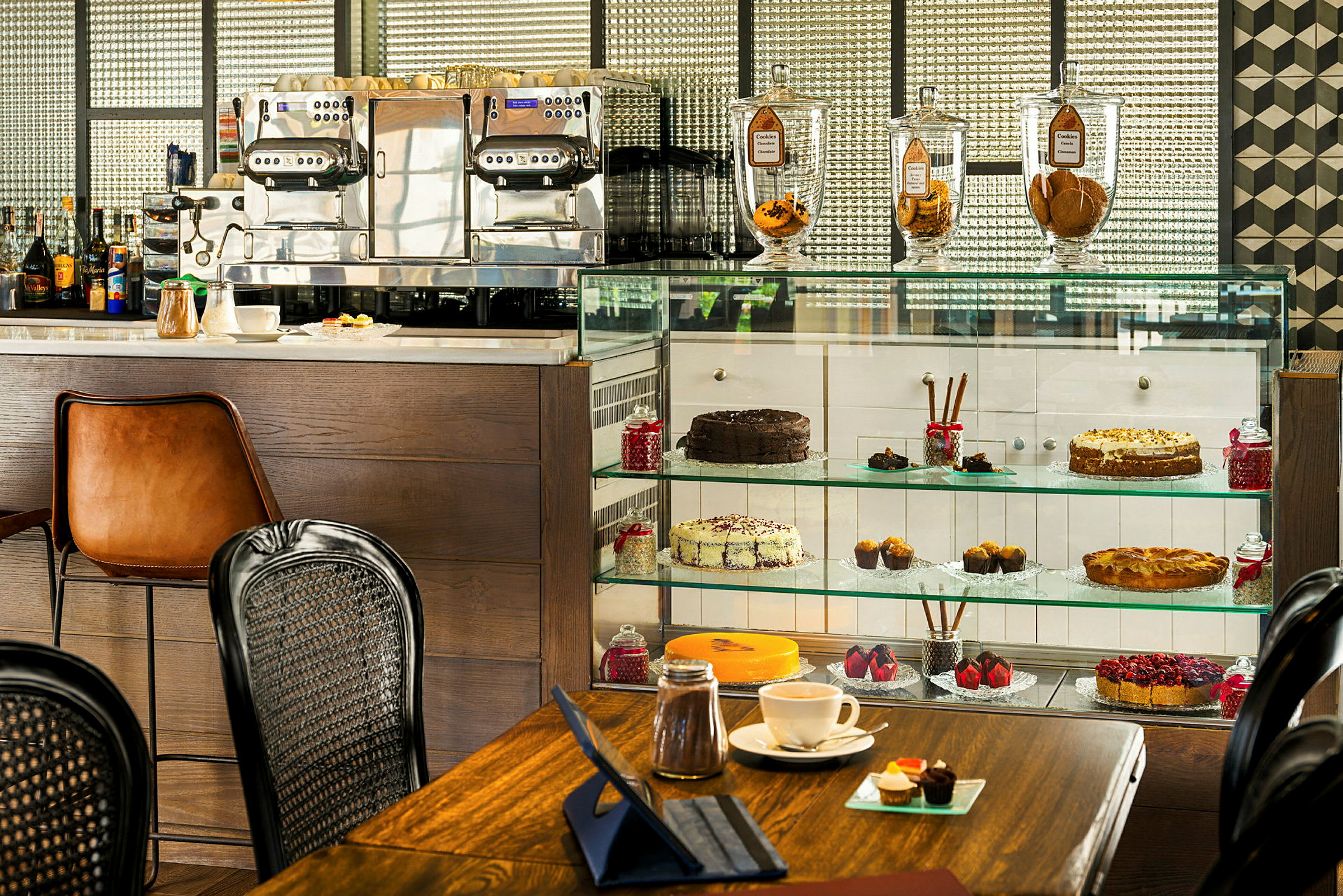 En bild av ett café med kakor och bakverk i en glasmonter, en espressomaskin och fina kaffekoppar på en bordsyta. (AI-genererad text)