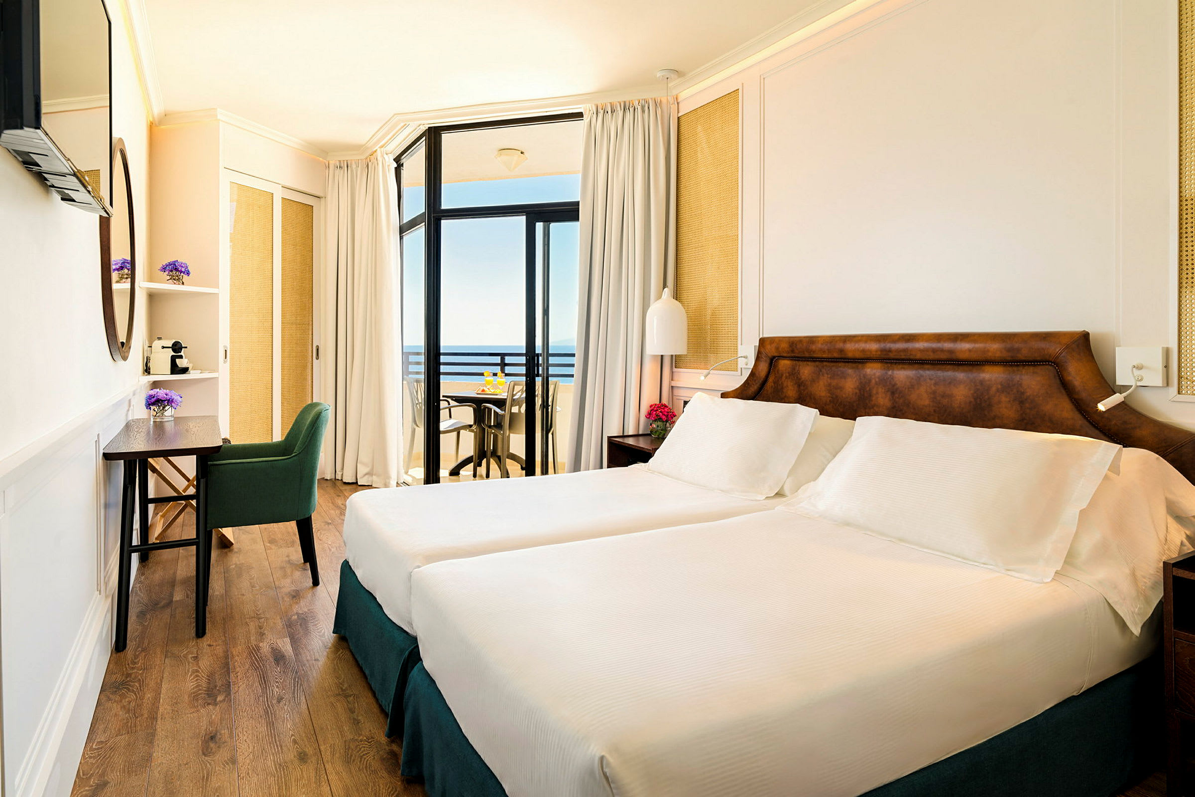 Ett hotellrum med två enkelsängar, ett skrivbord och en stol. Rummet har en balkong med utsikt mot havet. (AI-genererad text)