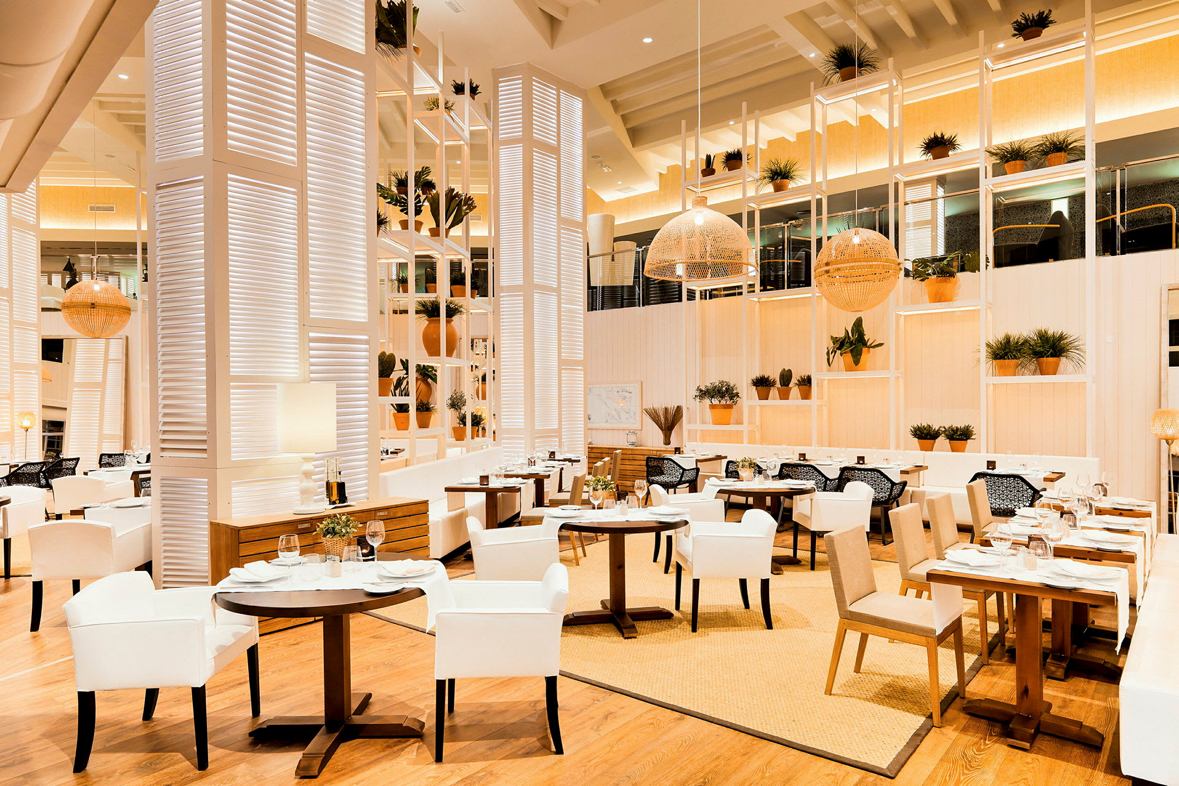 En elegant restaurang med ljus inredning, vita och beiga möbler samt trämaterial. Krukväxter pryder hyllor och borden. (AI-genererad text)