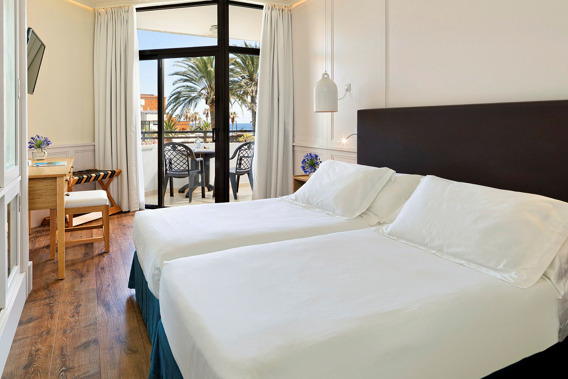 Ett ljust hotellrum med två enkelsängar, ett skrivbord och en balkong med utsikt över havet och palmer. (AI-genererad text)