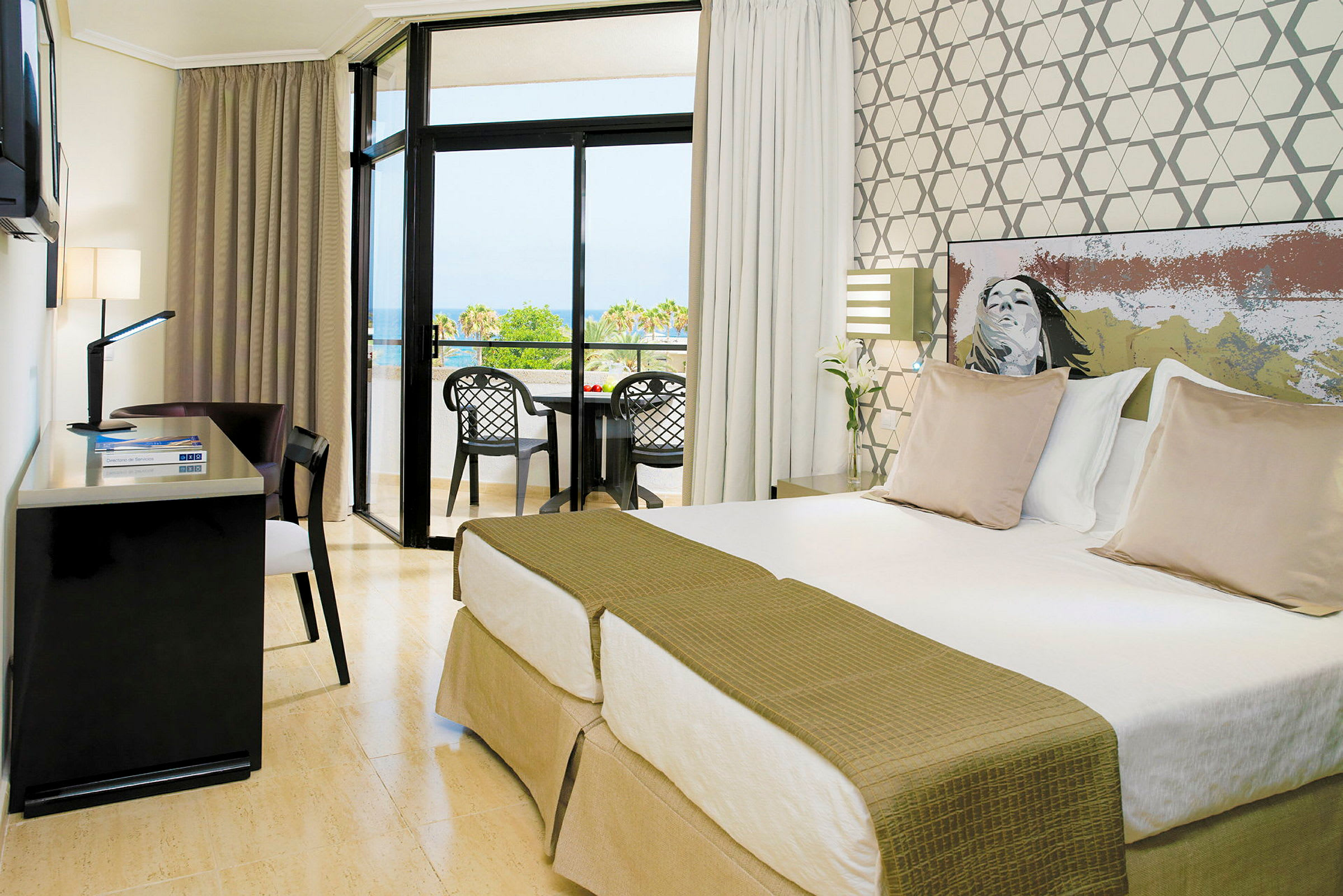 Ett hotellrum med två enkelsängar, ett skrivbord och en balkong med utsikt över ett grönområde och havet. (AI-genererad text)
