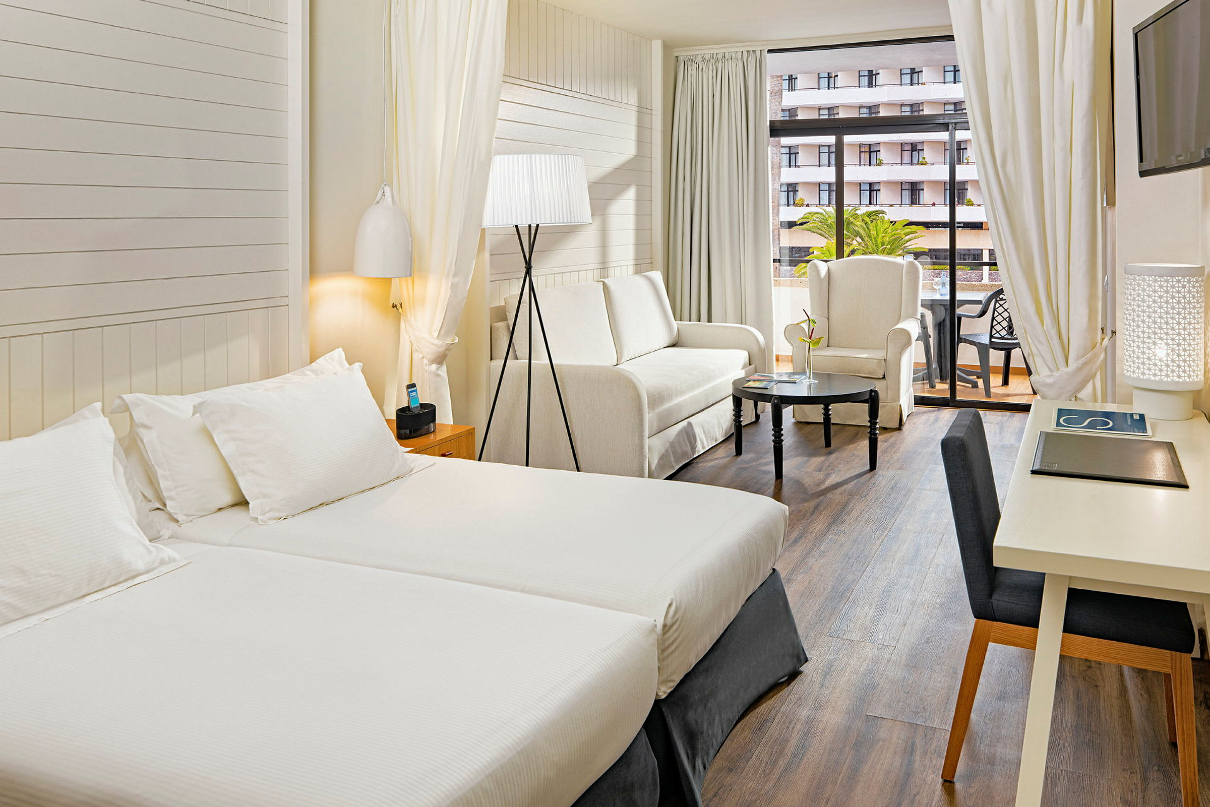 Ett hotellrum med två enkelsängar, en sittgrupp med soffa och fåtölj samt ett bord. Det finns även ett skrivbord och en balkong. (AI-genererad text)