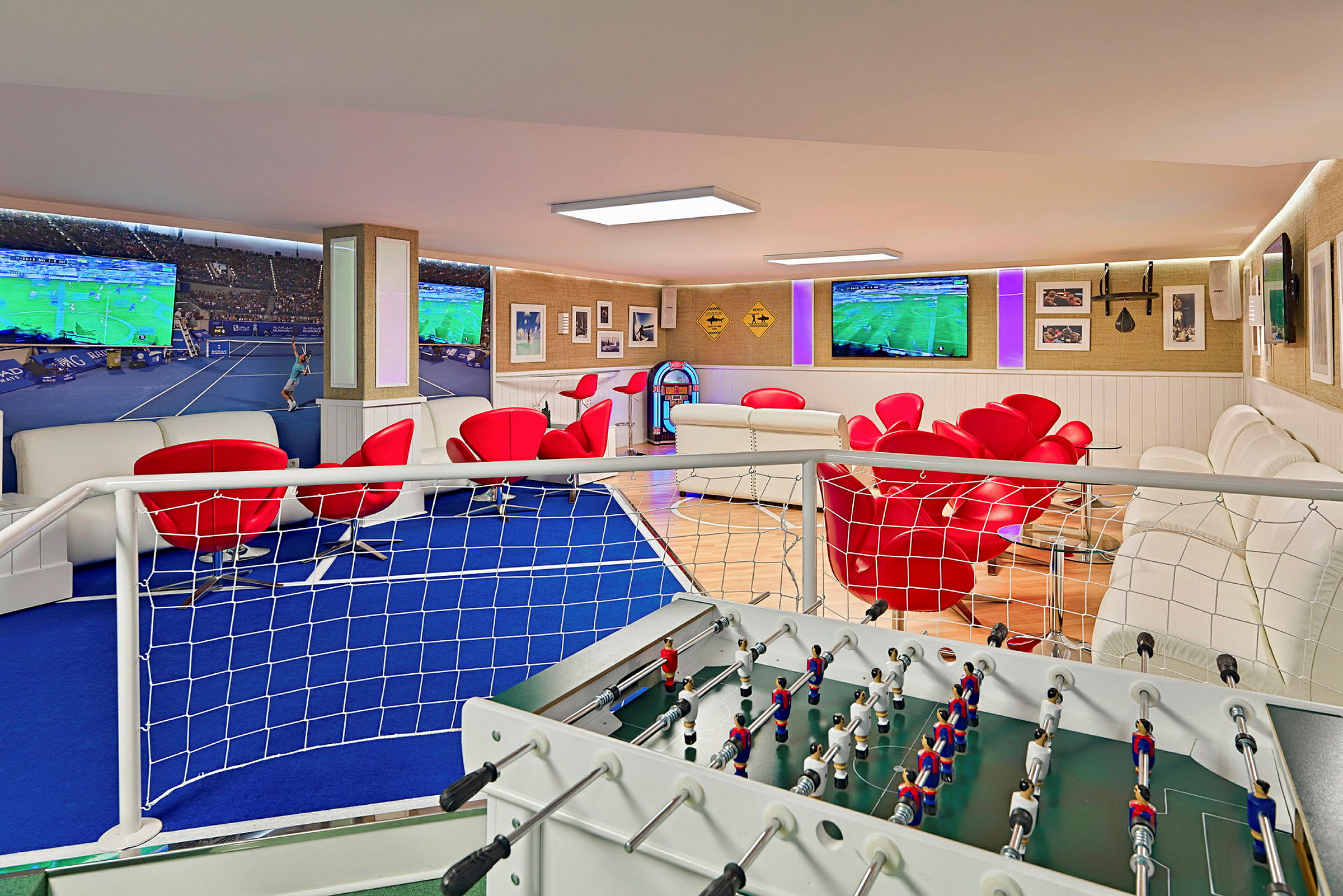 Ett rum med en foosballbord i förgrunden, röda stolar, vita soffor och flera stora TV-skärmar som visar sport. (AI-genererad text)