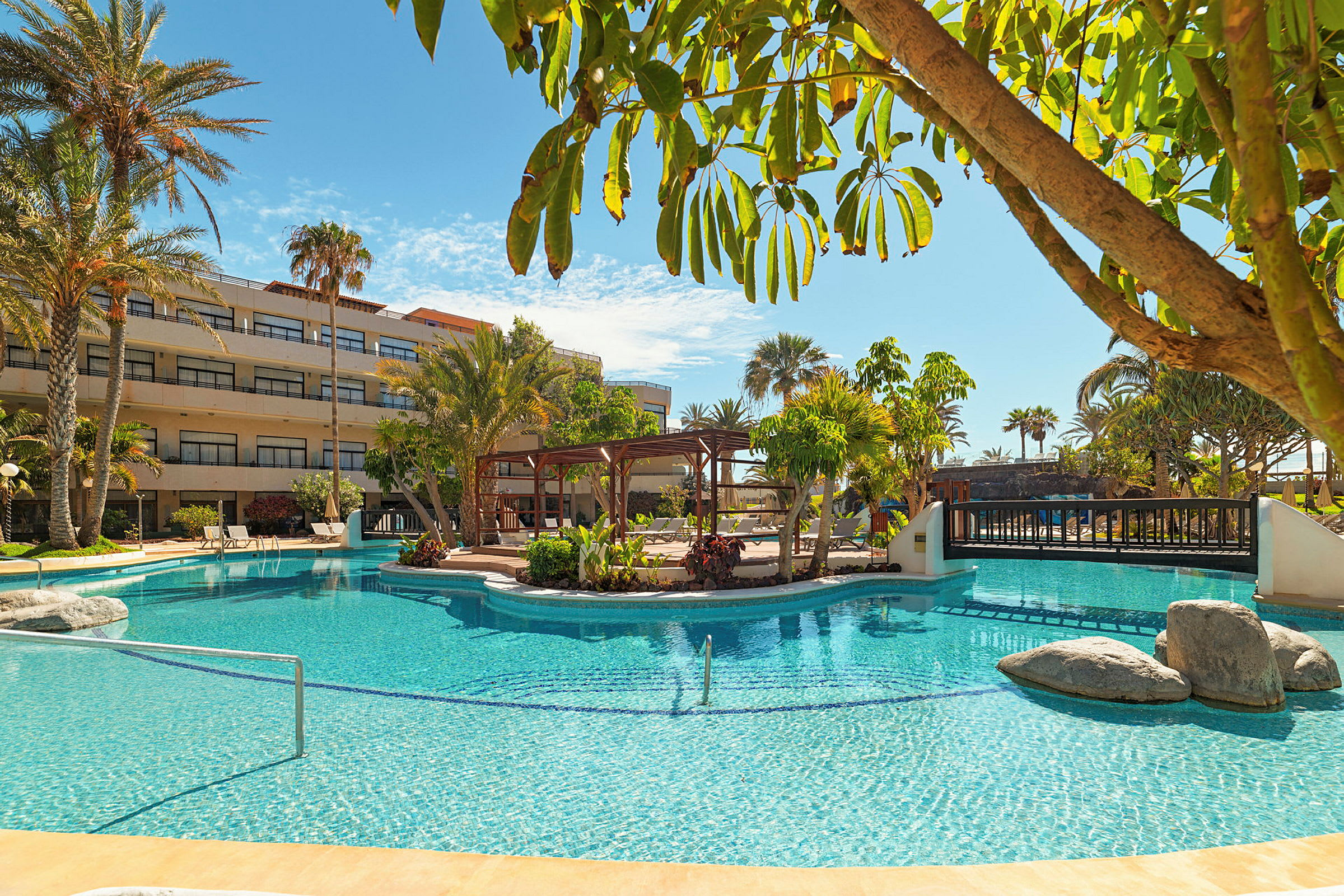 En pool vid ett hotell med omgivande grönska och palmer, samt byggnader i bakgrunden. (AI-genererad text)