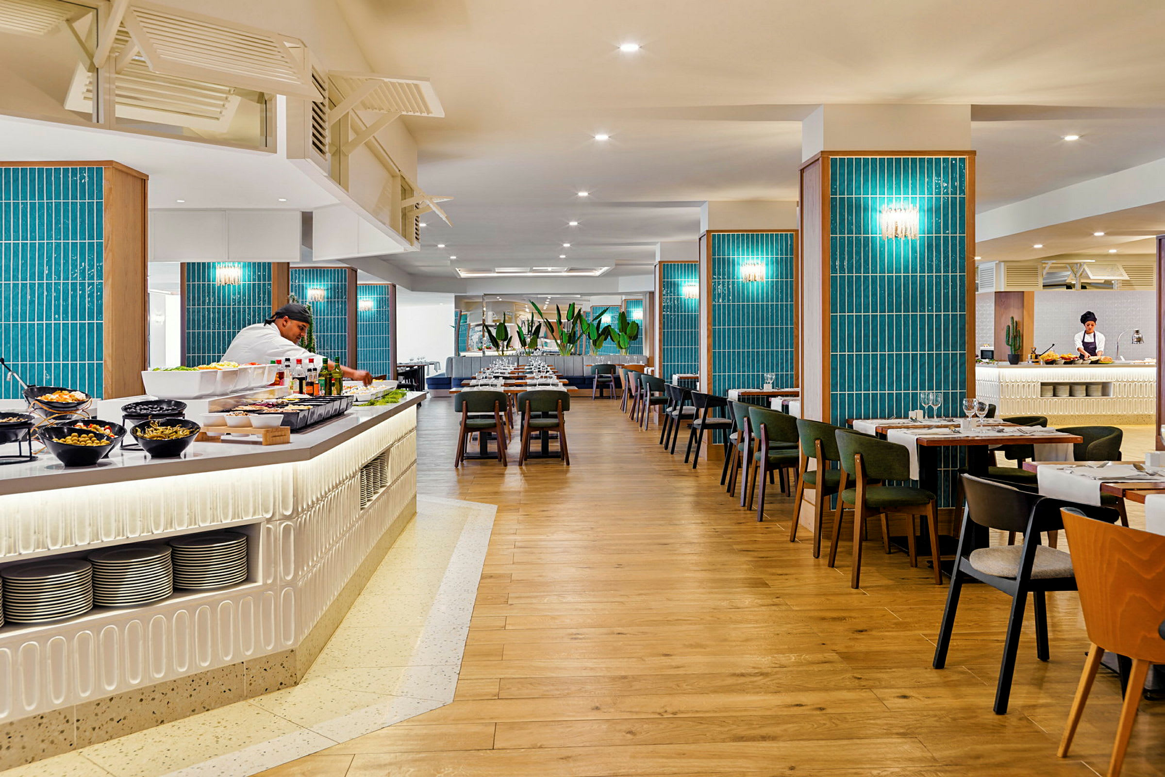 En rymlig restaurang med trägolv, blå väggdekorationer och bord med stolar. En buffetdisk med sallader och drycker syns i förgrunden, och en kock arbetar bakom disken. (AI-genererad text)