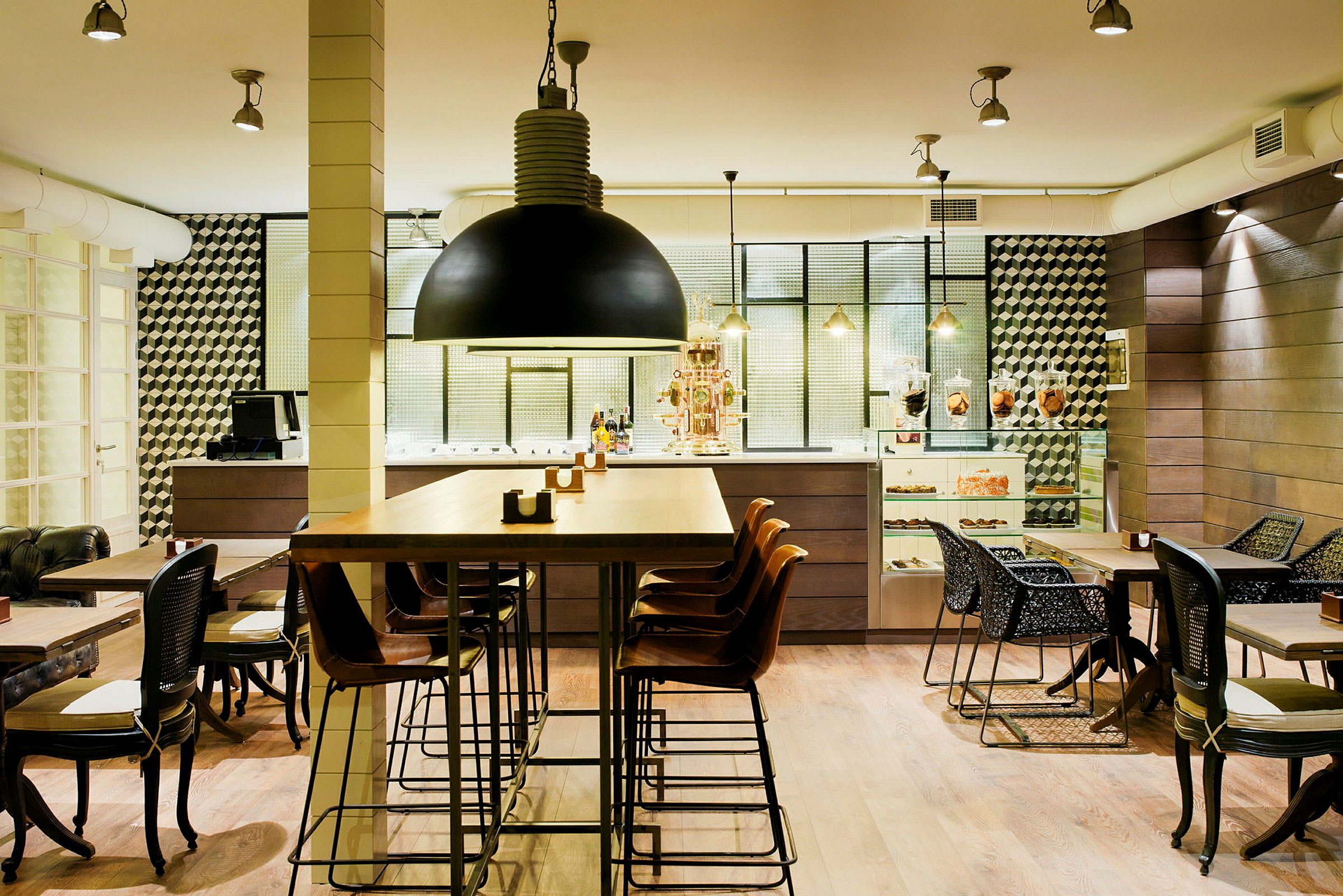 Ett modernt café med höga och låga bord, samt stolar i olika design. Inredningen är stilren med detaljer som svart hänglampa, geometriska väggmönster och ett skåp med bakverk. (AI-genererad text)