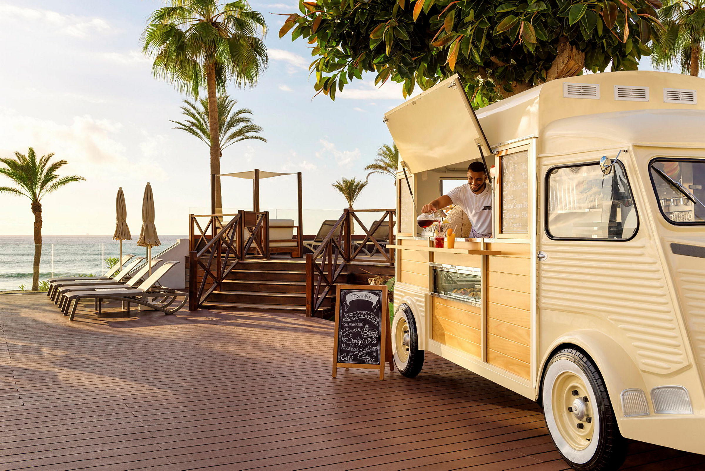 En strandnära plats med solstolar, parasoller, träd och en food truck. (AI-genererad text)