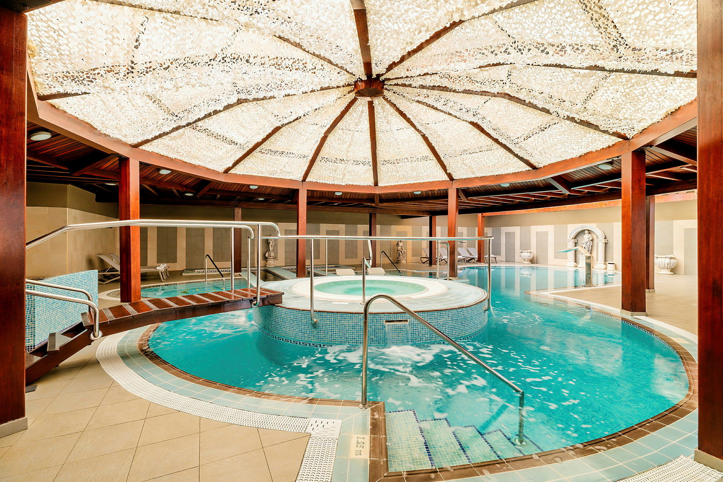 Interiör av ett spa med en pool och jacuzzi under ett dekorerat innertak. Träbjälkar och områden för avkoppling är synliga. (AI-genererad text)