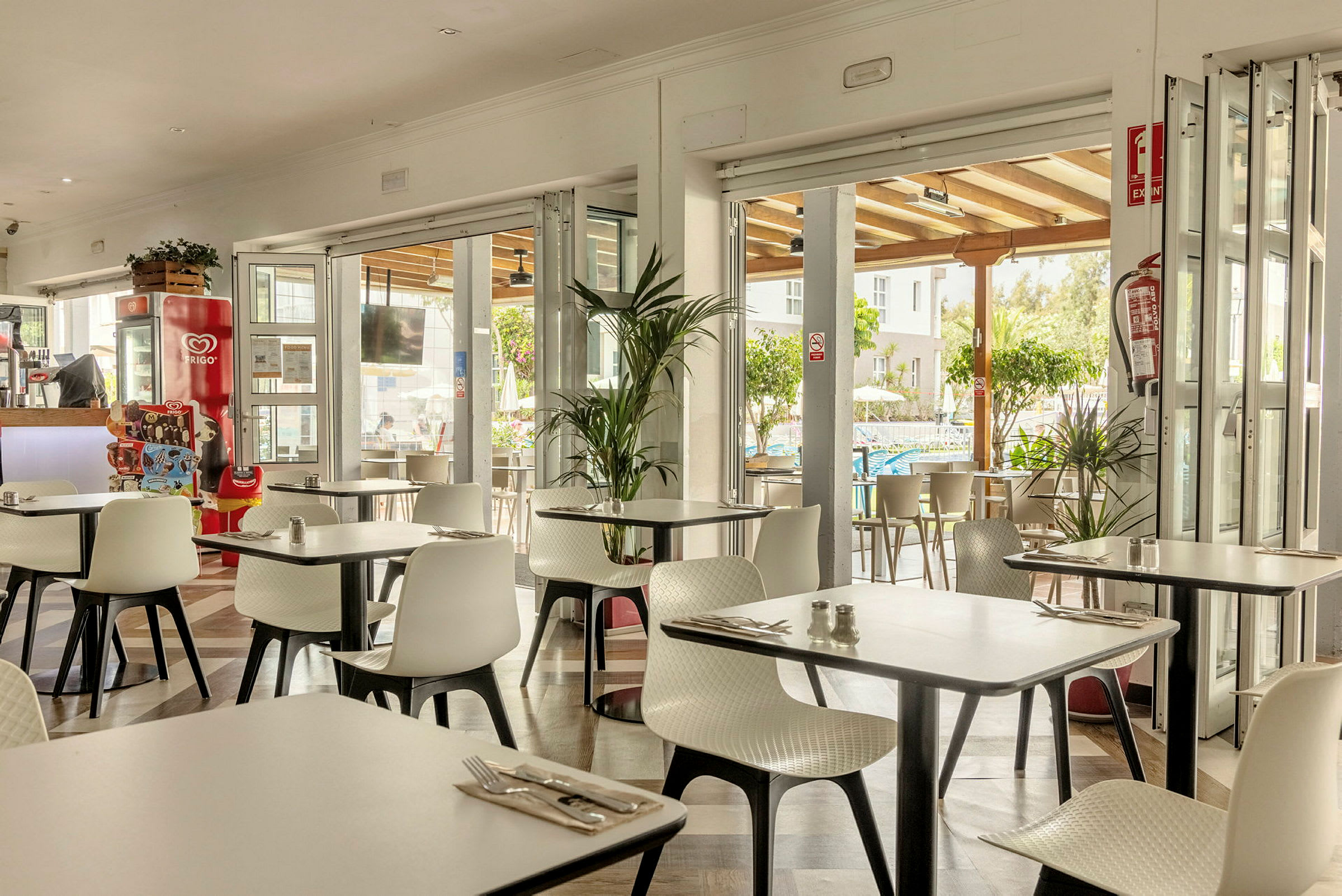En restaurang med vita stolar och bord, öppen mot en terrass och omgivande grönska. (AI-genererad text)