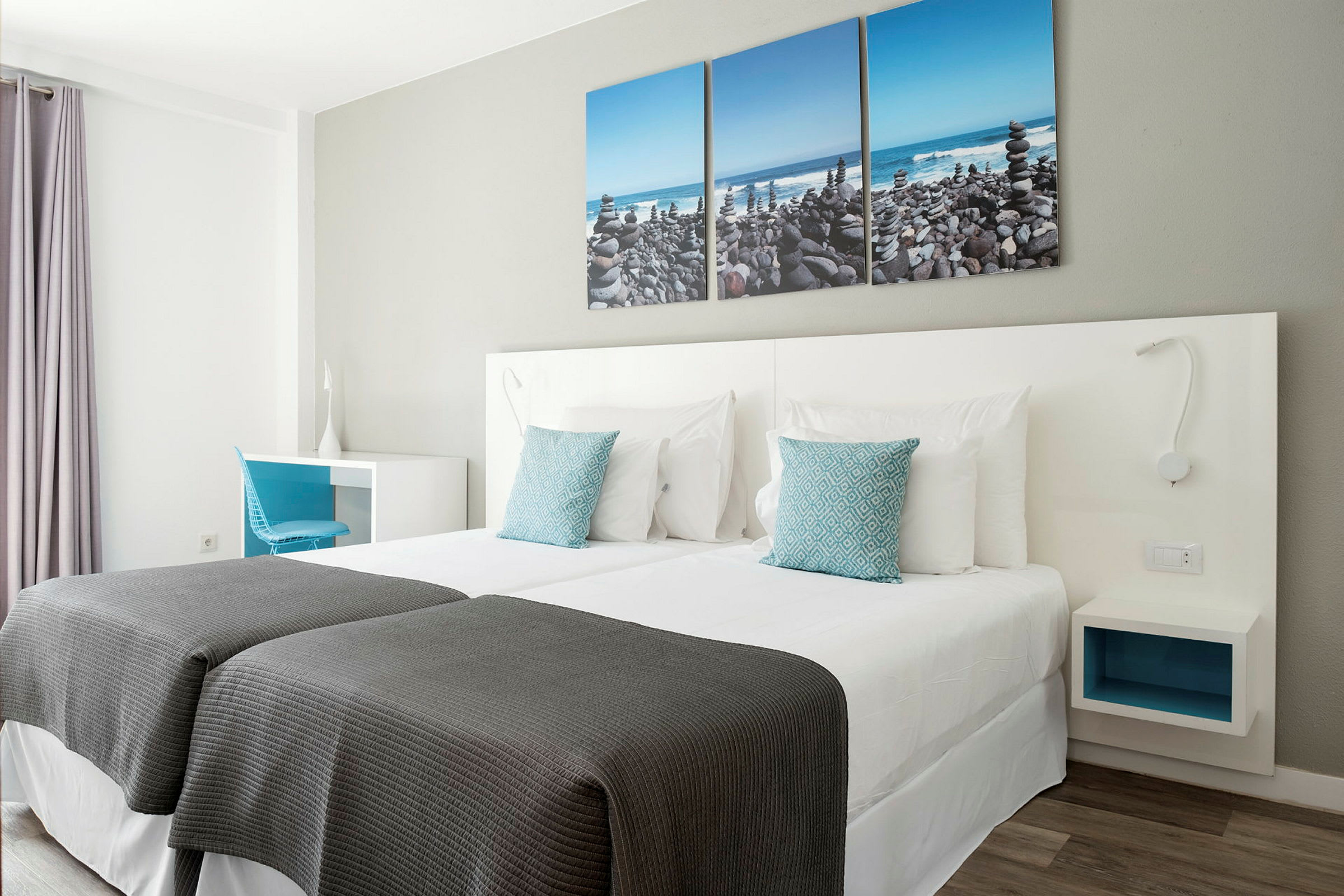Ett hotellrum med två enkelsängar, vita sängkläder och blå prydnadskuddar. På väggen hänger en tredelad tavla föreställande en stenstrand. (AI-genererad text)