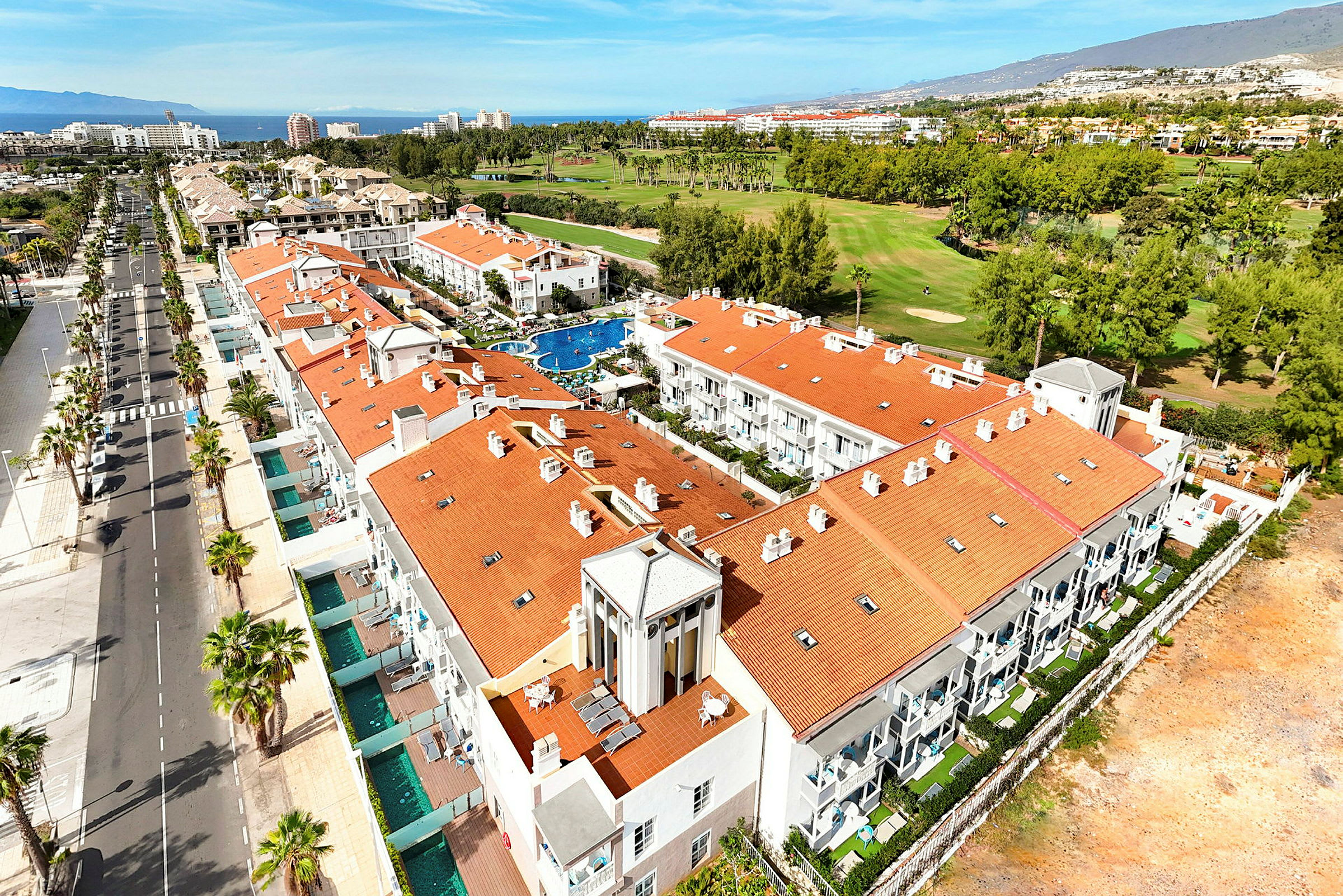 Flygfoto av ett hotellområde med röda tak, en pool, träd och en golfbana i bakgrunden. (AI-genererad text)