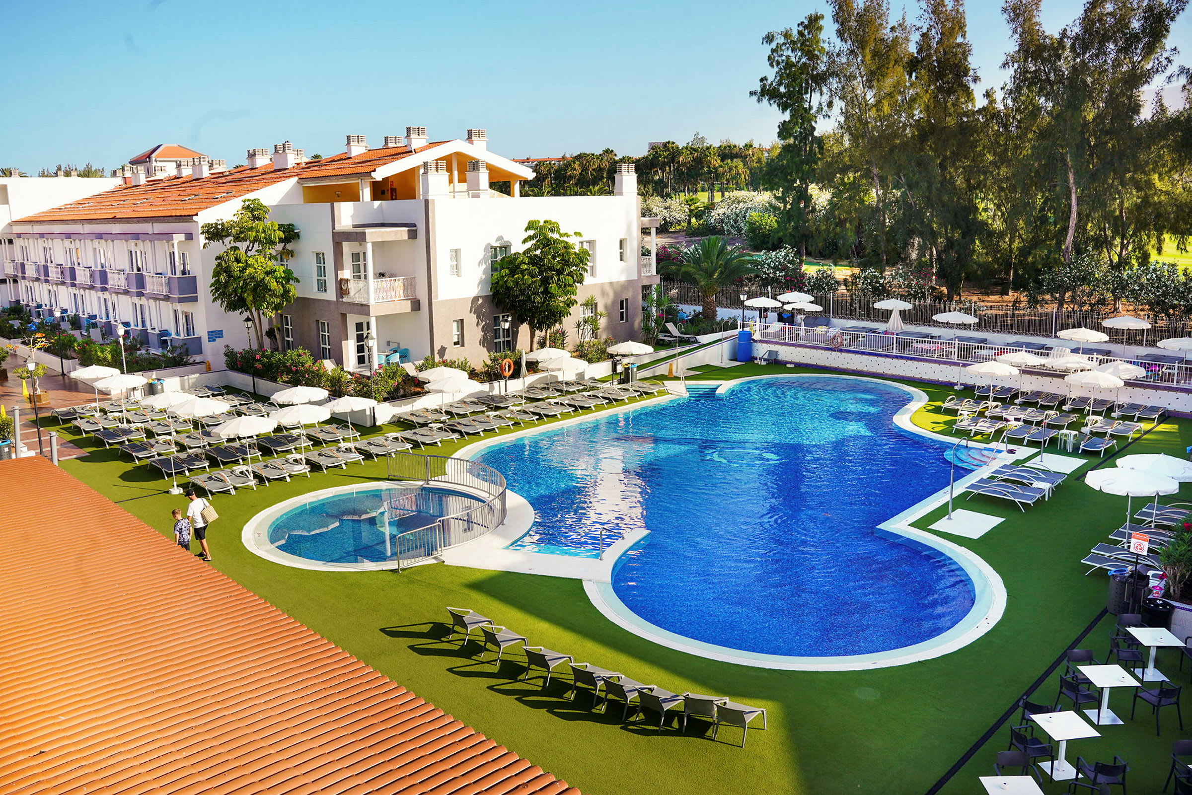 Bild av en hotellpool med solstolar och parasoller. Byggnader och grönska syns i bakgrunden. (AI-genererad text)