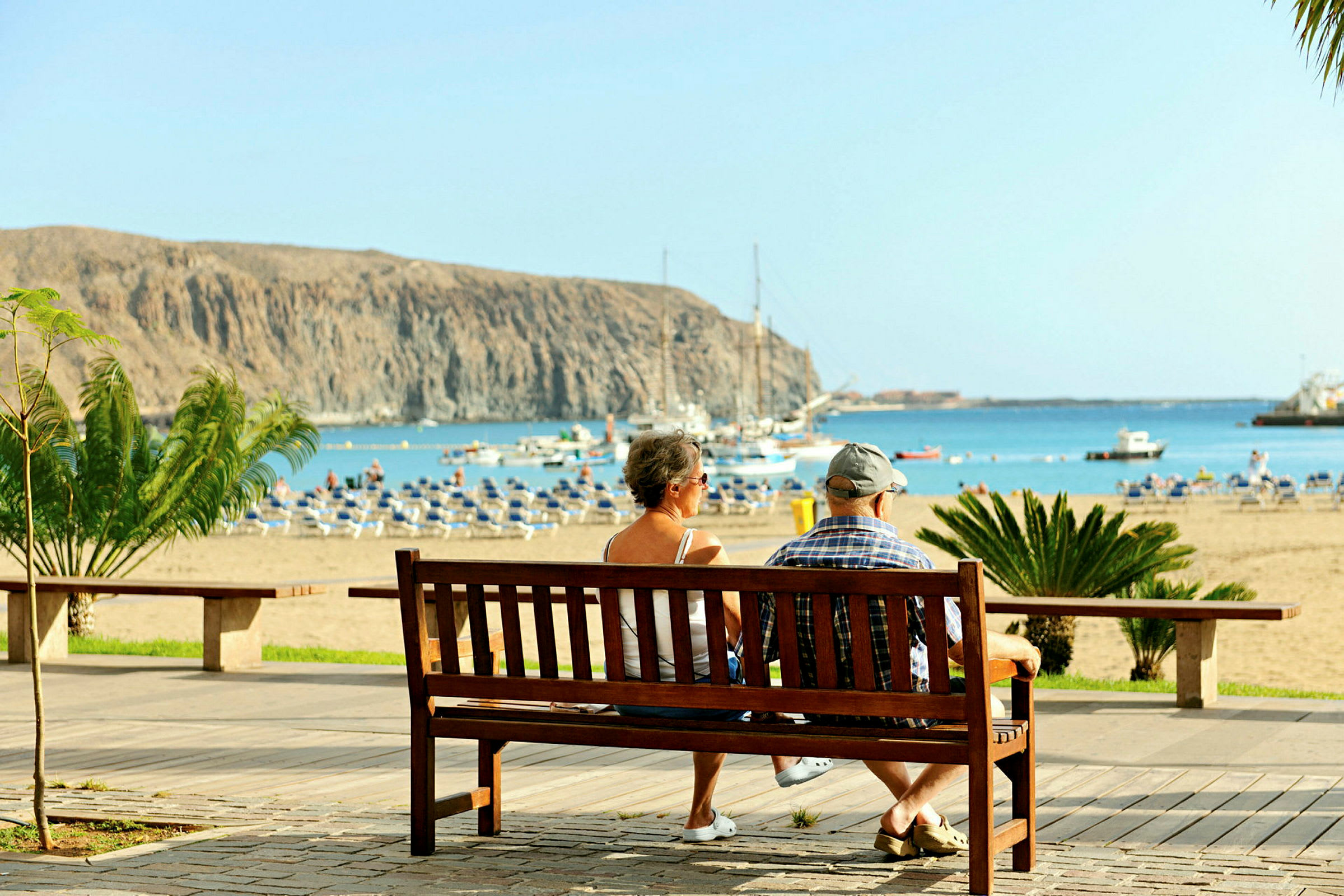 Två personer sitter på en bänk och ser ut över en sandstrand och havet med båtar och klippor i bakgrunden. (AI-genererad text)