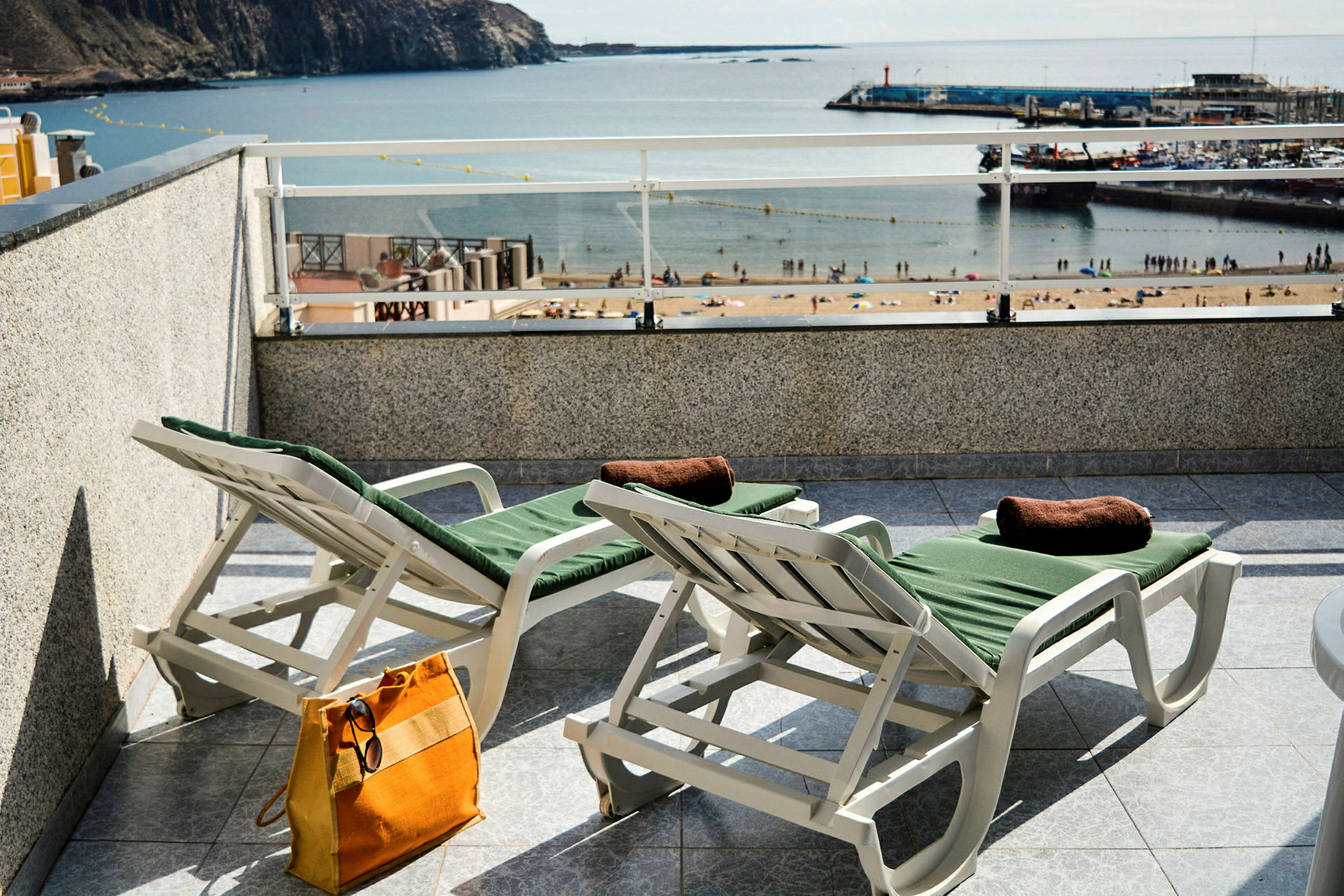 Två solstolar med gröna dynor på en terrass med utsikt över en strand och hamn. (AI-genererad text)