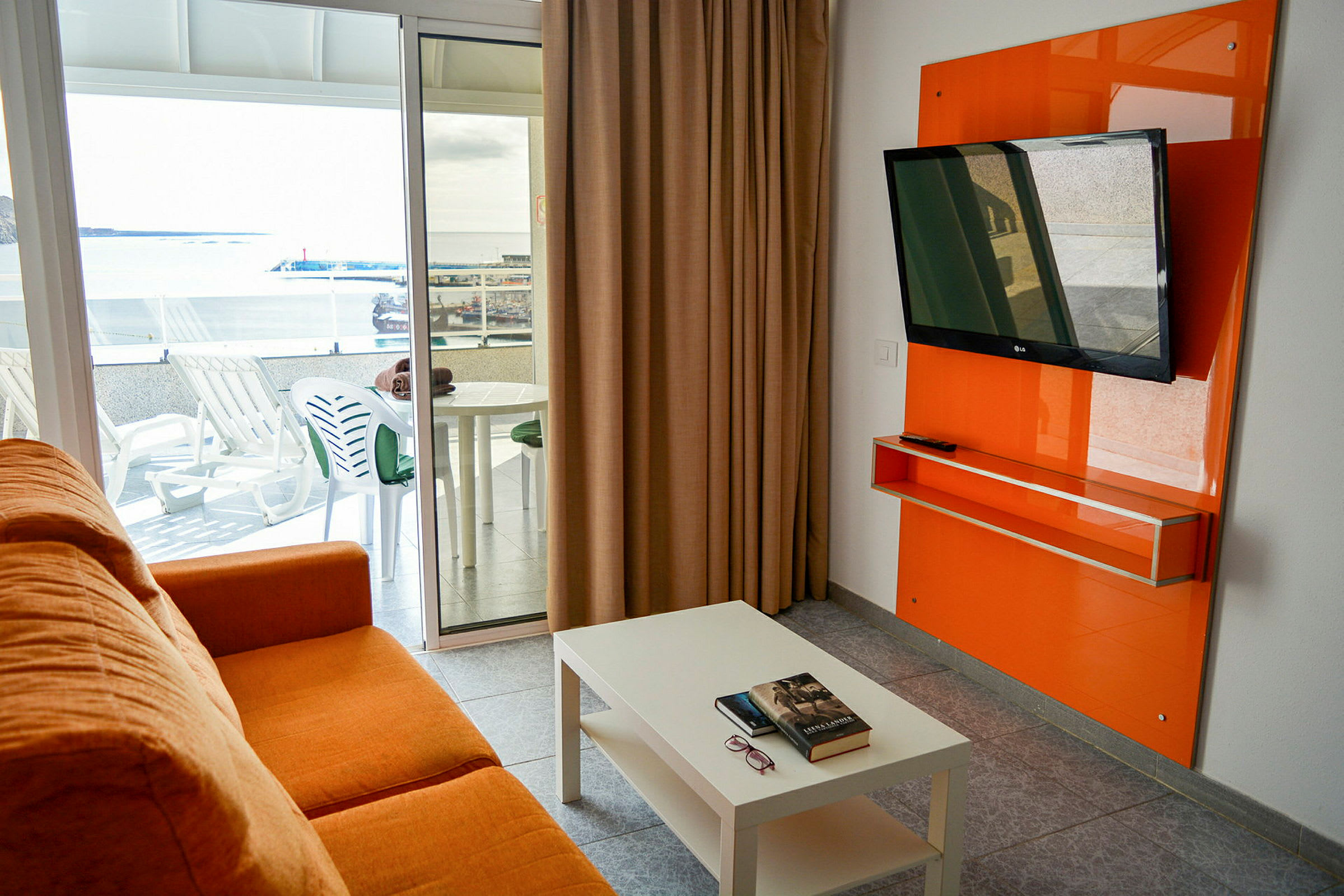 En orange soffa i ett rum med ett vitt bord, en platt-TV på väggen och en balkong med utemöbler samt utsikt över ett hamnområde. (AI-genererad text)