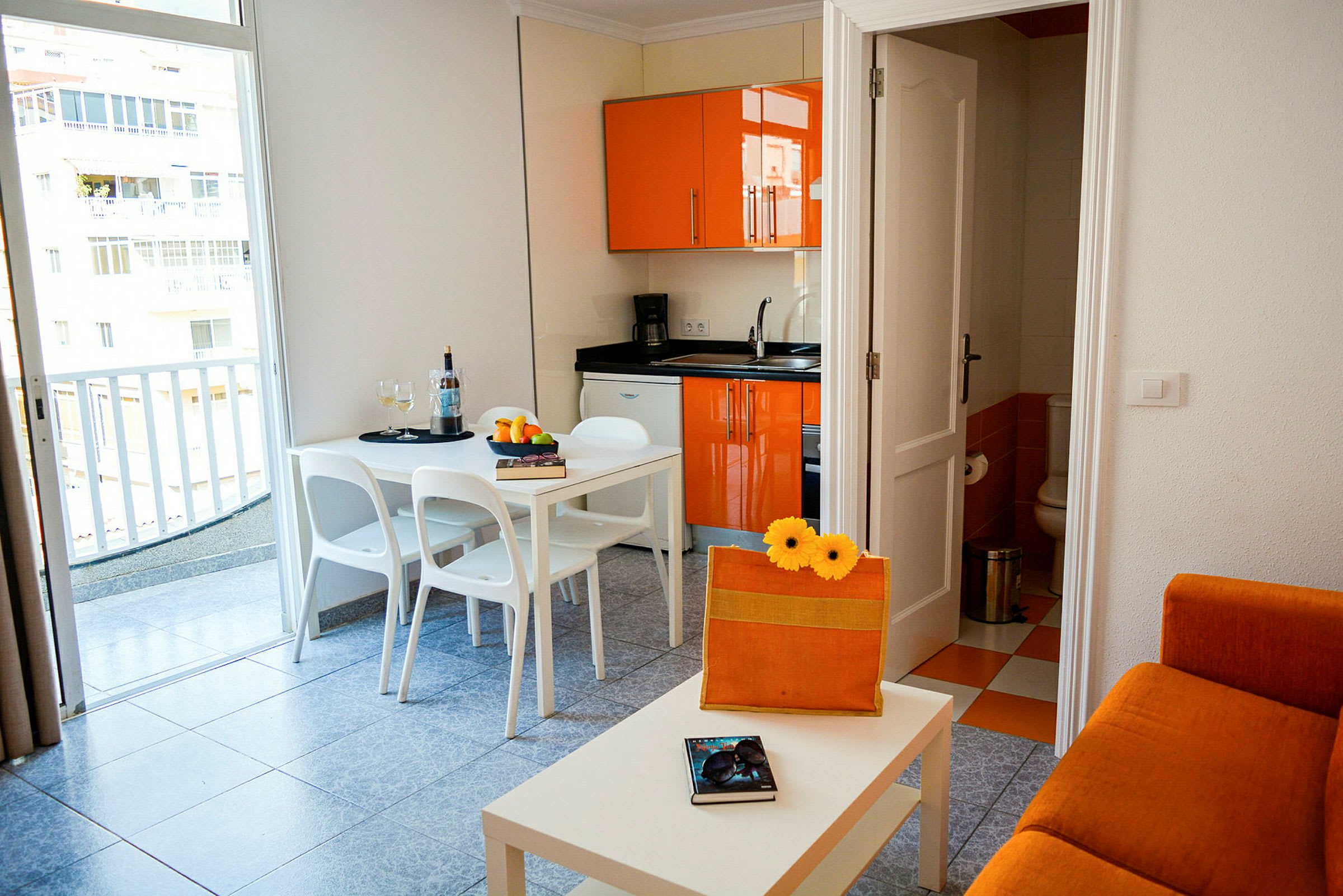 En liten lägenhet med orange köksinredning, en vit matplats och en soffa. (AI-genererad text)