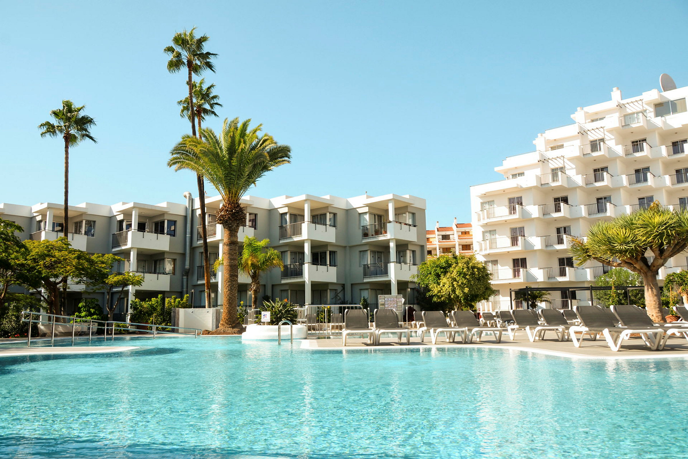 Bild på en pool med solstolar framför flerfamiljshus och palmer. (AI-genererad text)