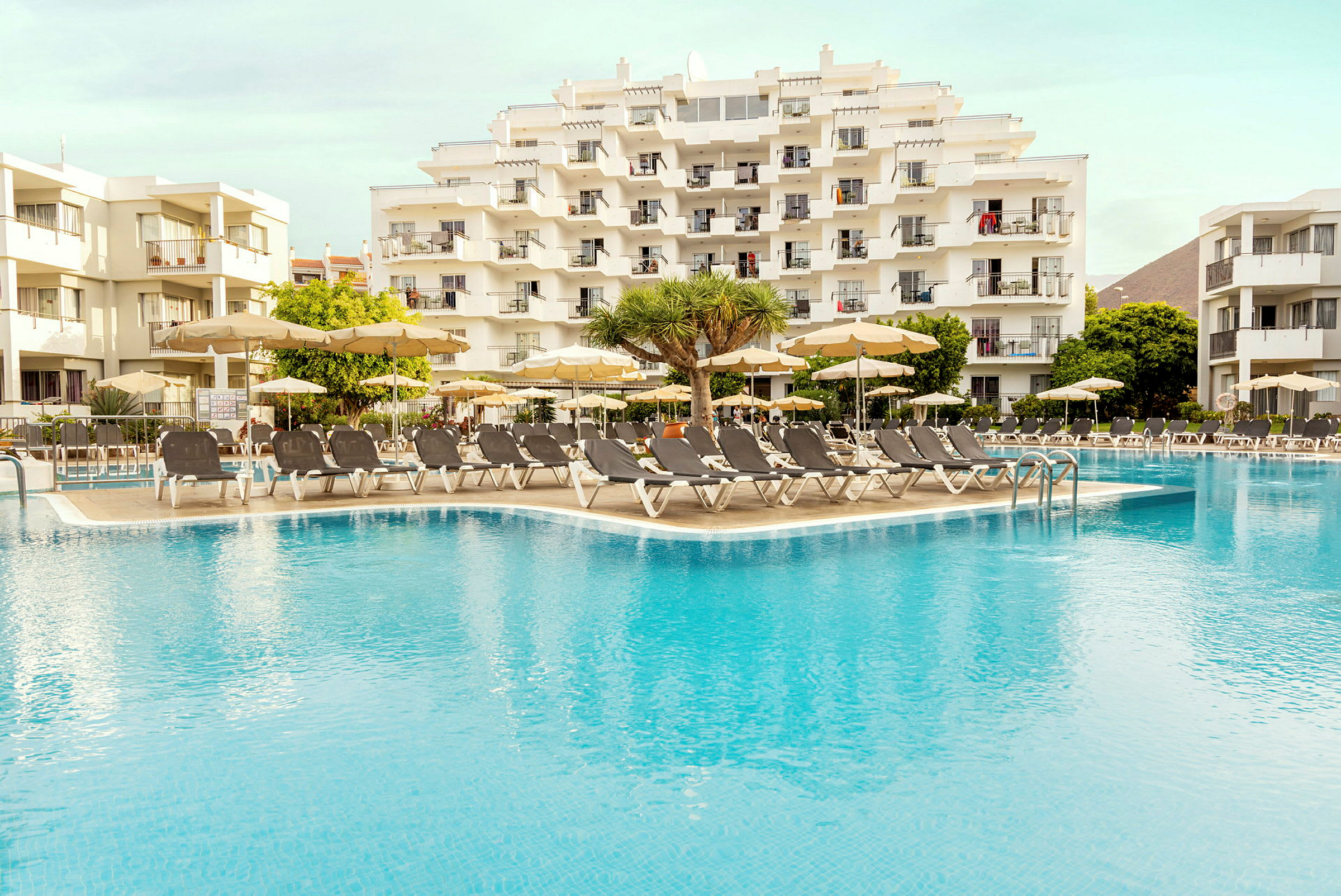 Bild av ett hotellkomplex med solstolar och parasoll vid en swimmingpool. (AI-genererad text)