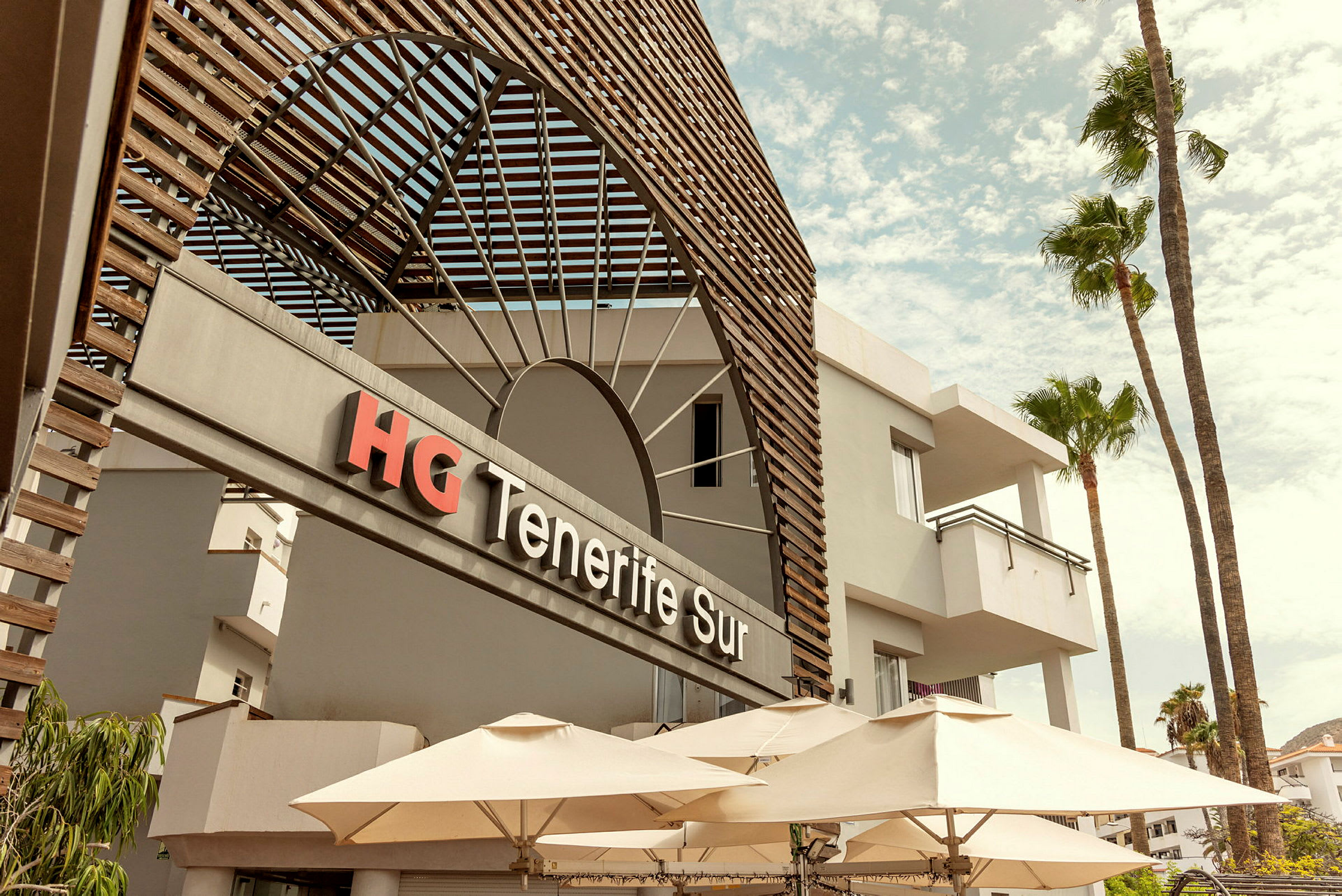 Framsidan av HG Tenerife Sur-hotellet med parasoller och palmer. (AI-genererad text)