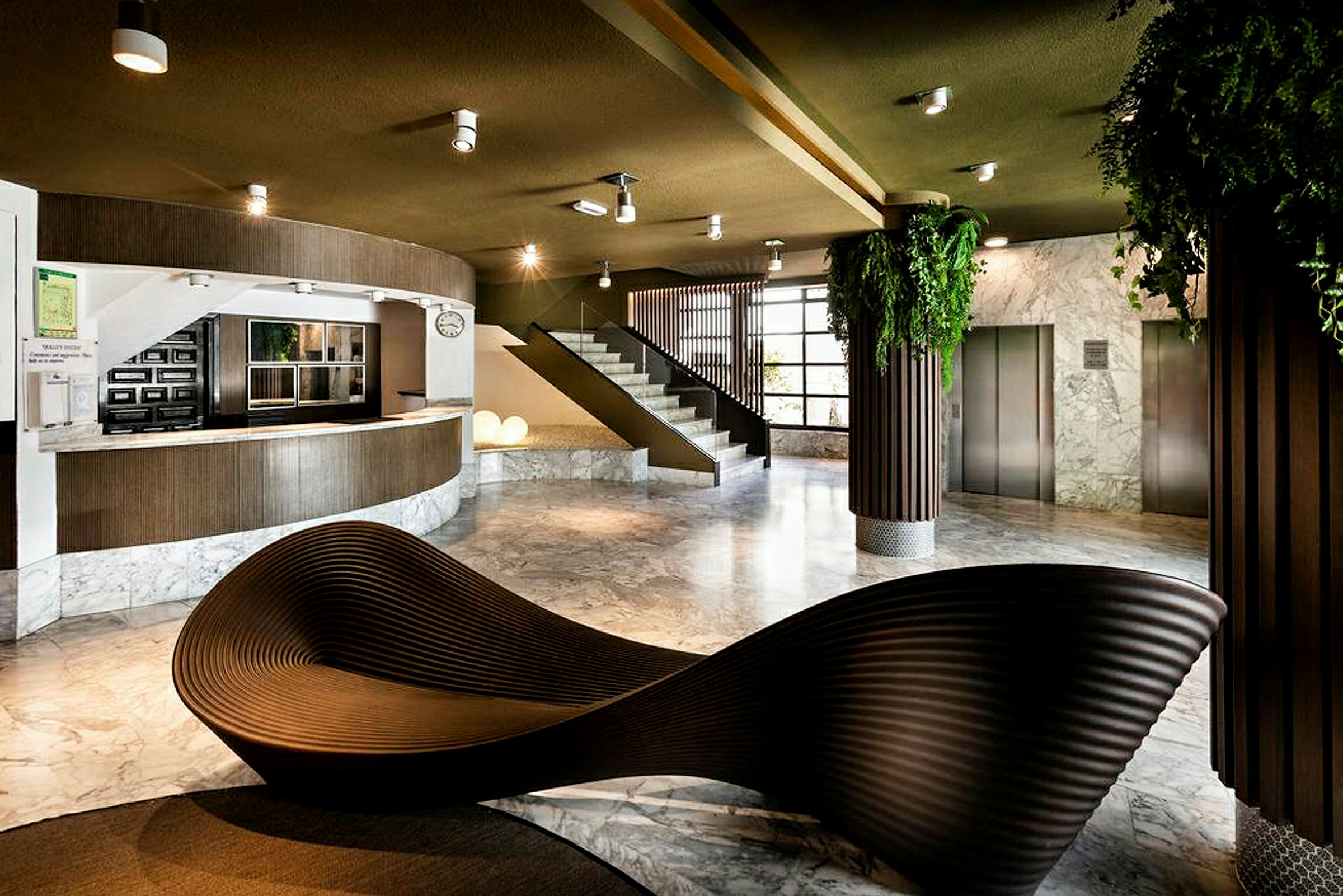 Ett modernt lobbymiljö med receptionsdisk, trappa och hissar. Inredningen inkluderar en skulptural sittmöbel och gröna växter. (AI-genererad text)