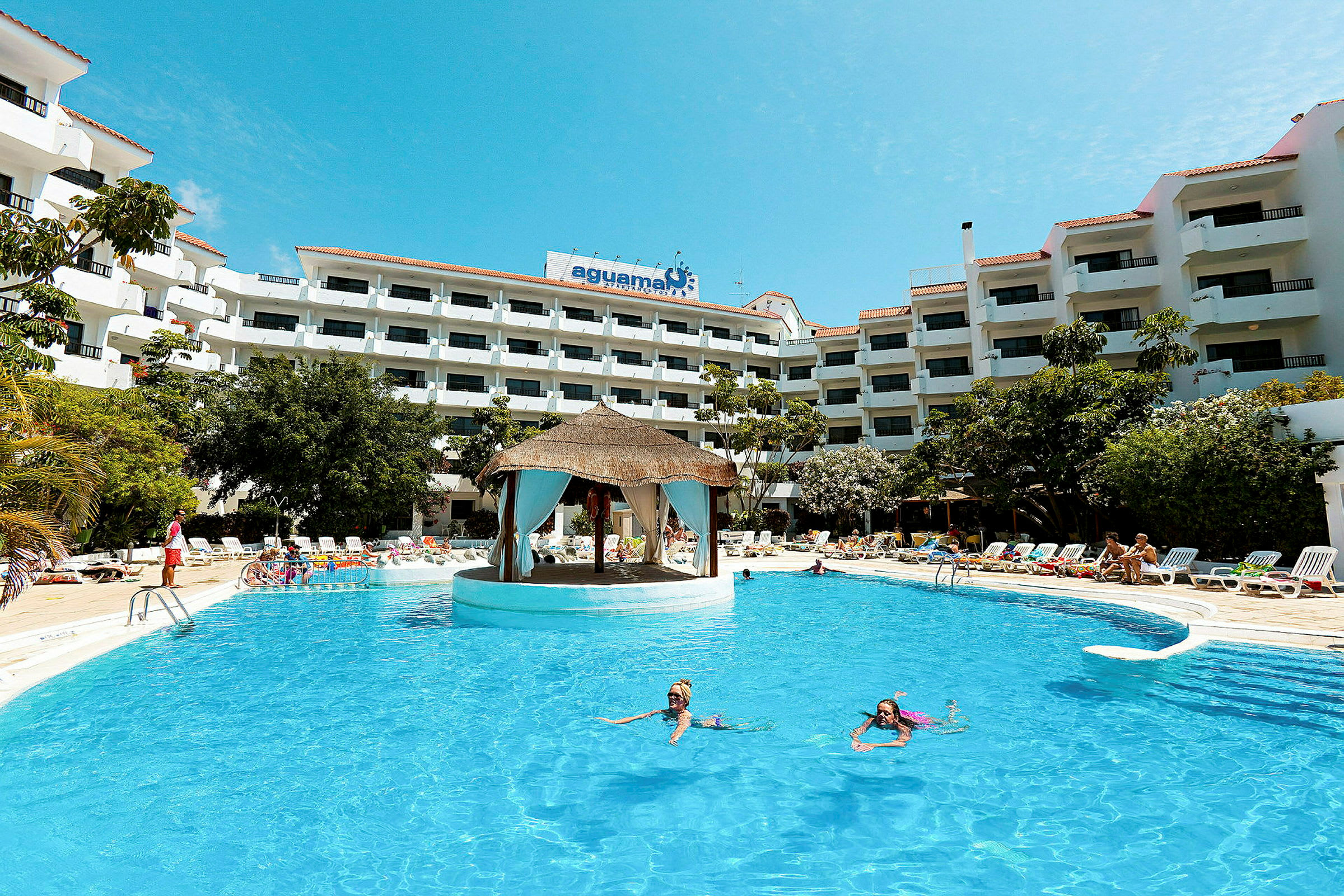 En stor pool med simmande människor framför en hotellbyggnad med flera våningar. Byggnaden omges av träd och solsängar. (AI-genererad text)