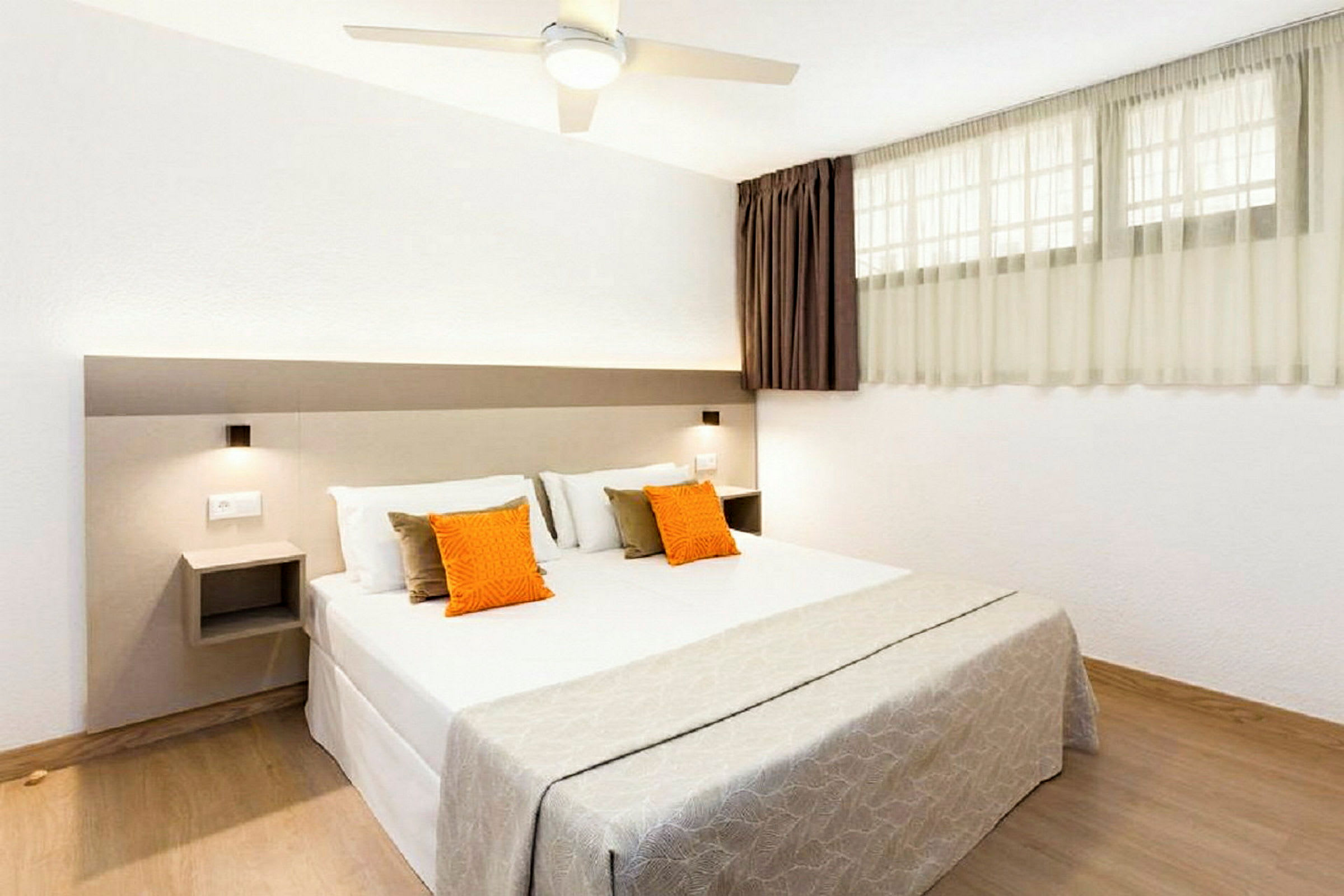 Ett sovrum med en dubbelsäng som har vita lakan och två dekorativa kuddar i orange och beige. Rummet är ljust med en fönstervägg och neutrala färger. (AI-genererad text)