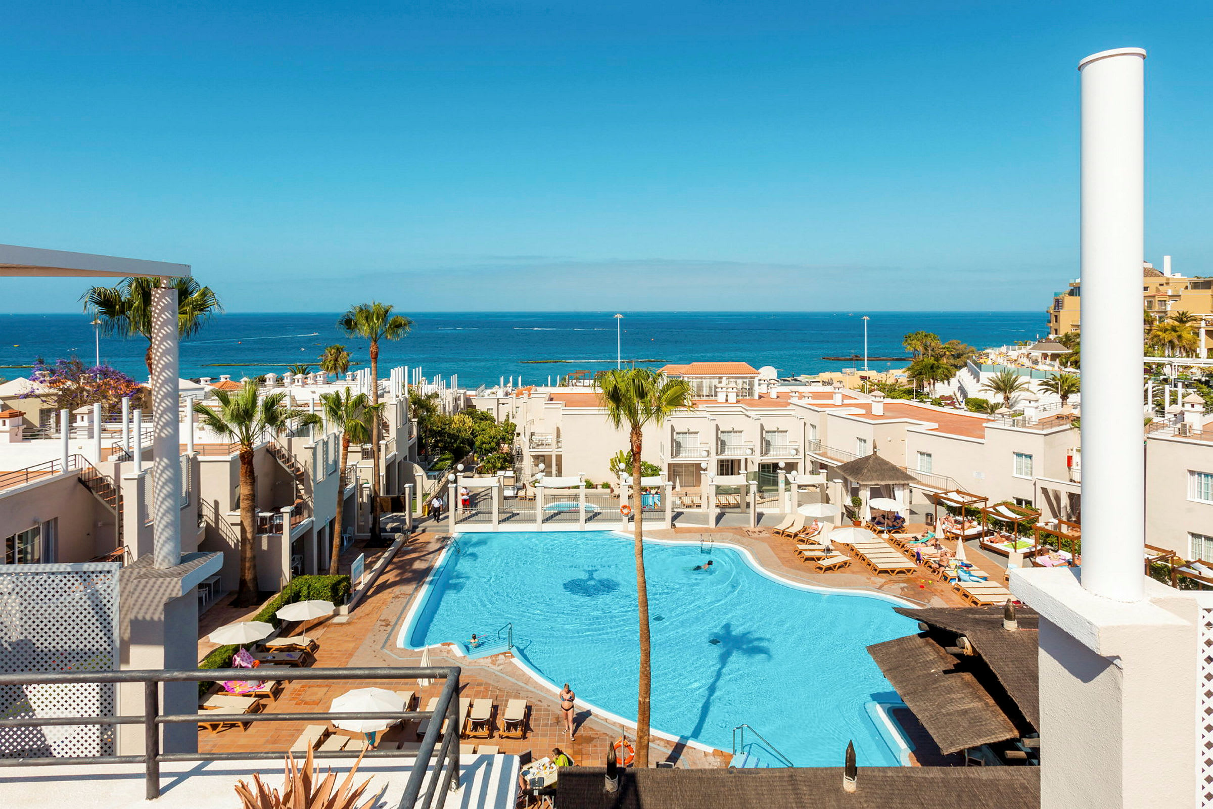 Bild av en semesteranläggning med pool, solstolar och palmträd, med utsikt över havet. (AI-genererad text)