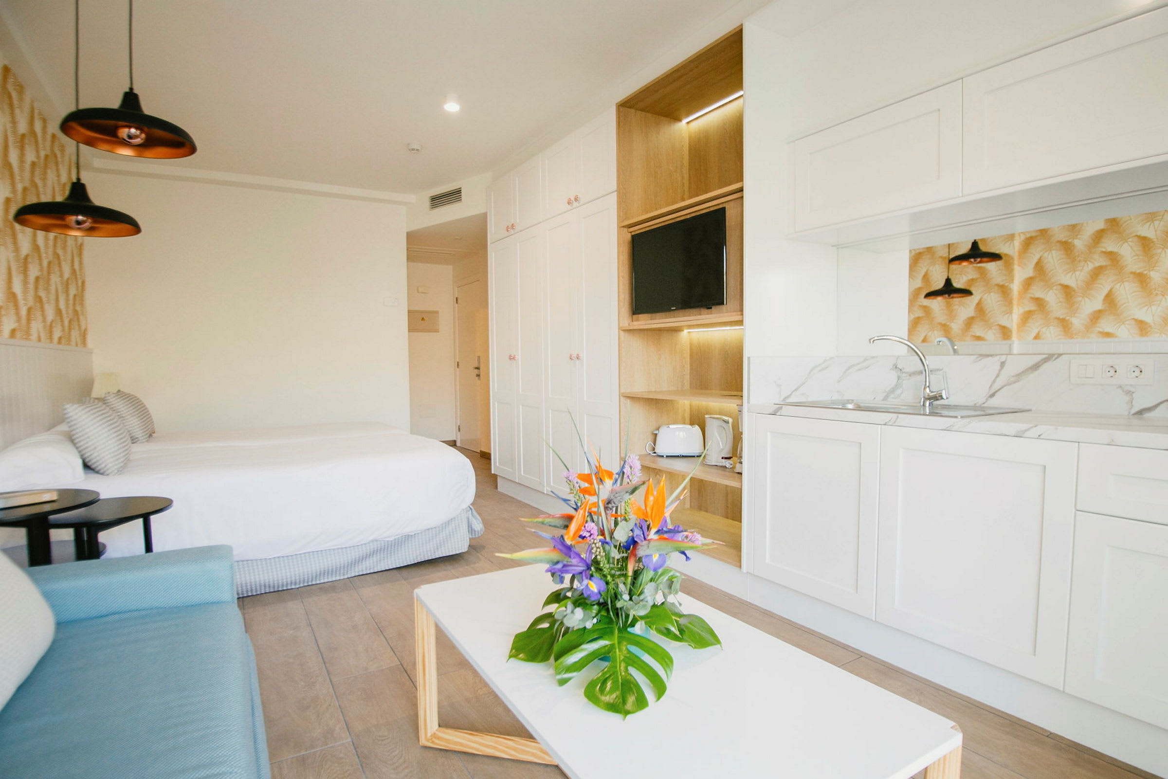 Ett modernt hotellrum med en dubbelsäng, soffa, litet kök och ett bord med en blombukett. (AI-genererad text)