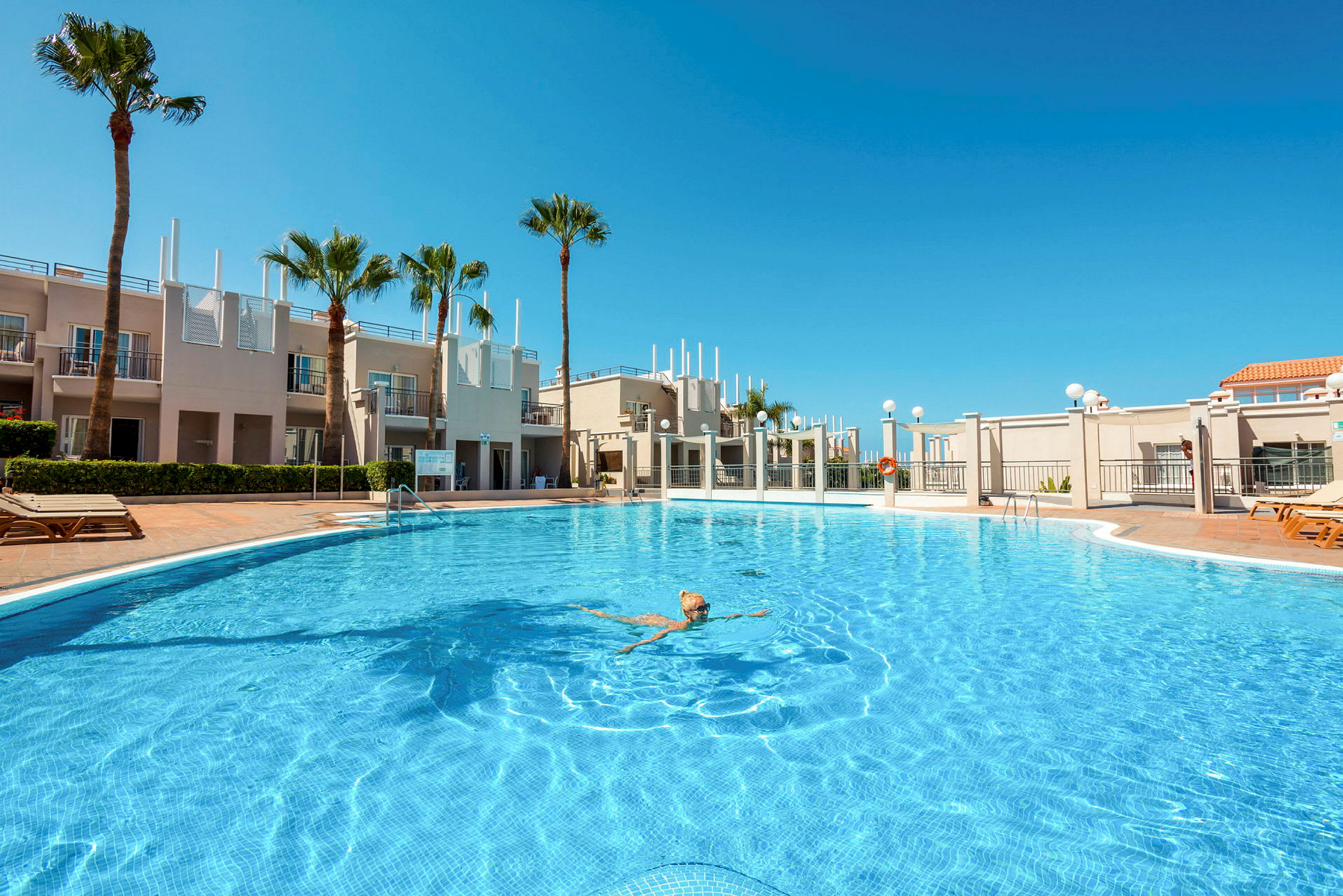 Person simmar i en stor swimmingpool omgiven av byggnader och palmer. (AI-genererad text)