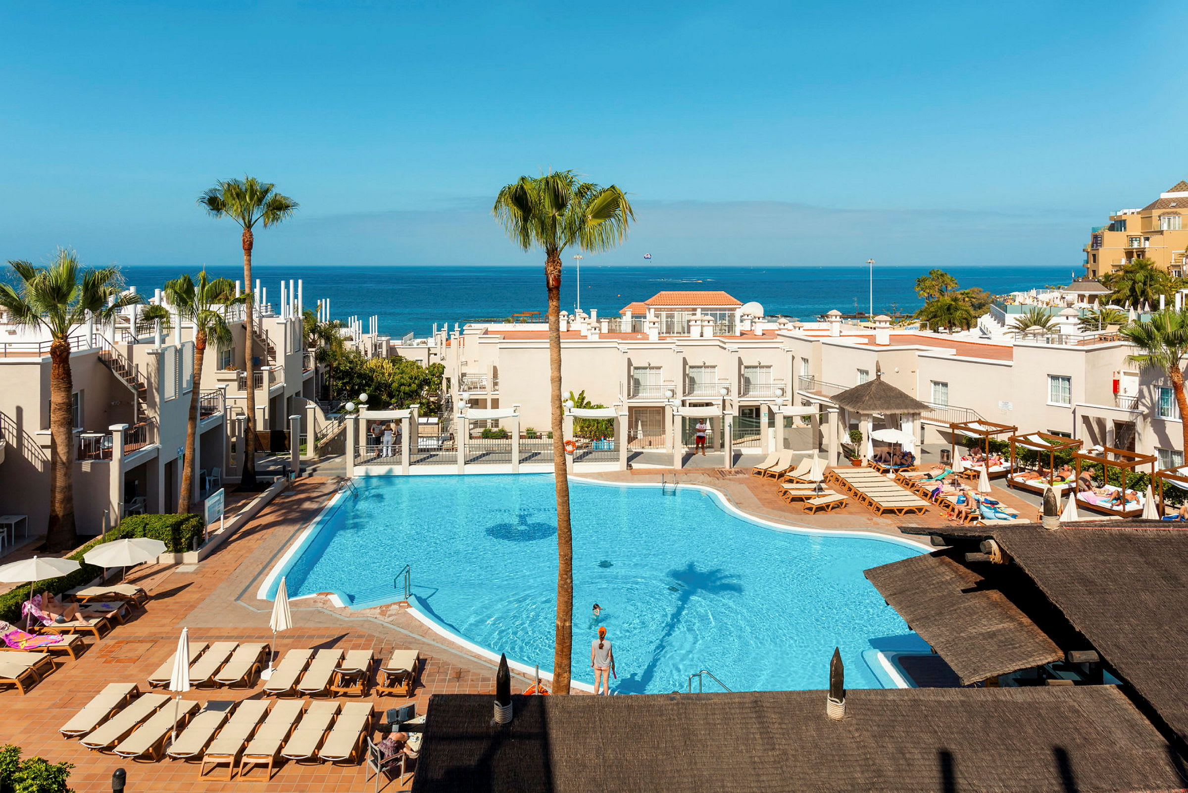 Vy över ett hotellområde med pool och solstolar, havet synligt i bakgrunden. (AI-genererad text)