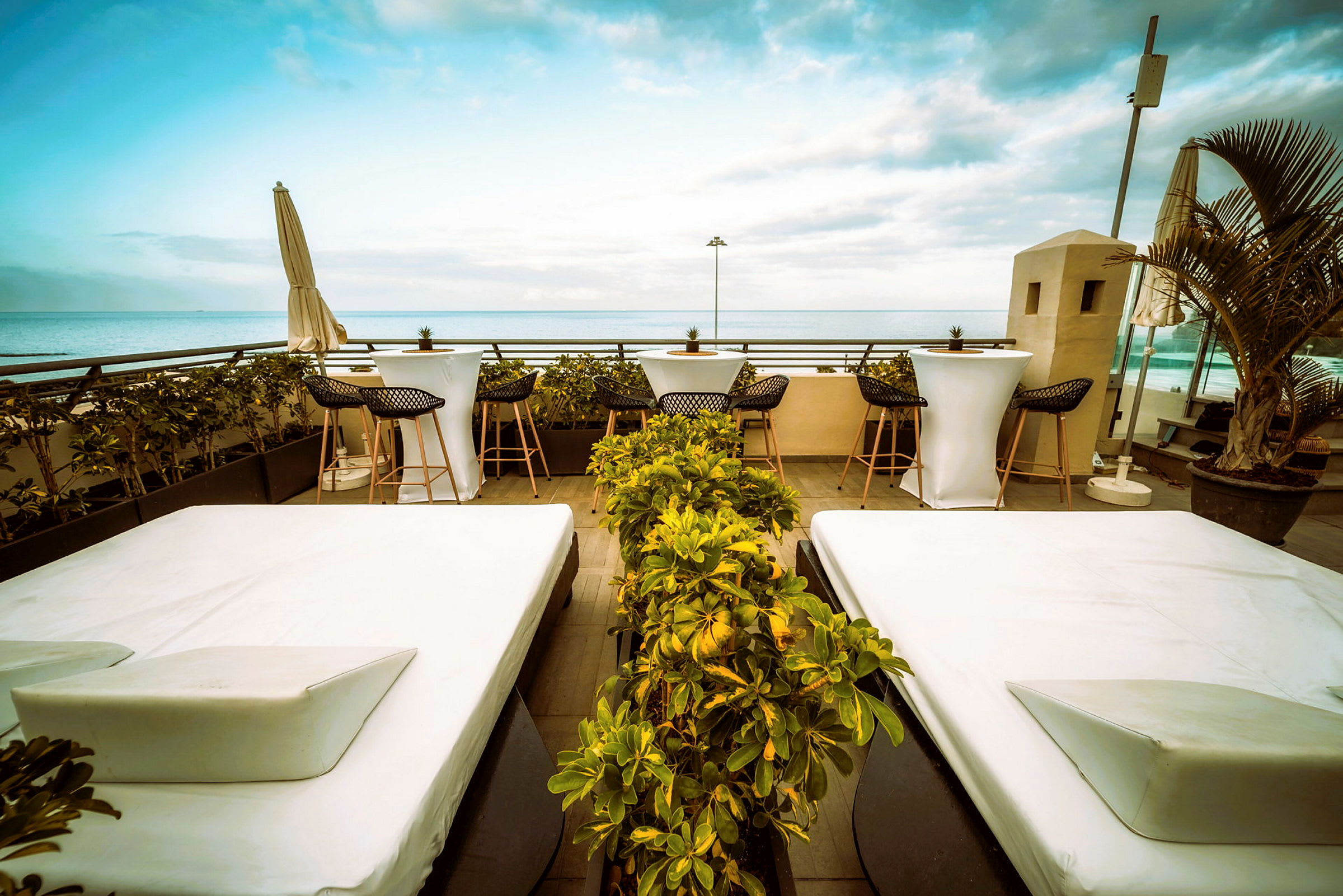 En terrass med vita solsängar och små bord med stolar. I bakgrunden syns havet och blå himmel. (AI-genererad text)