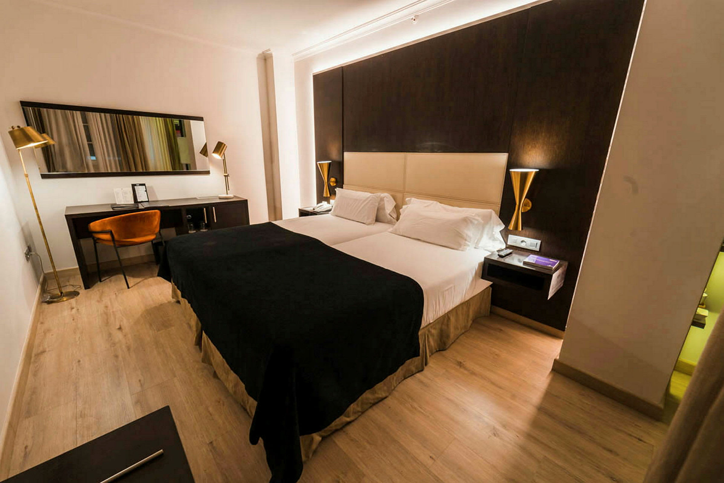 Ett hotellrum med en dubbelsäng, mörka och ljusa färgtoner på möbler samt en skrivbordshörna med stol och spegel. (AI-genererad text)