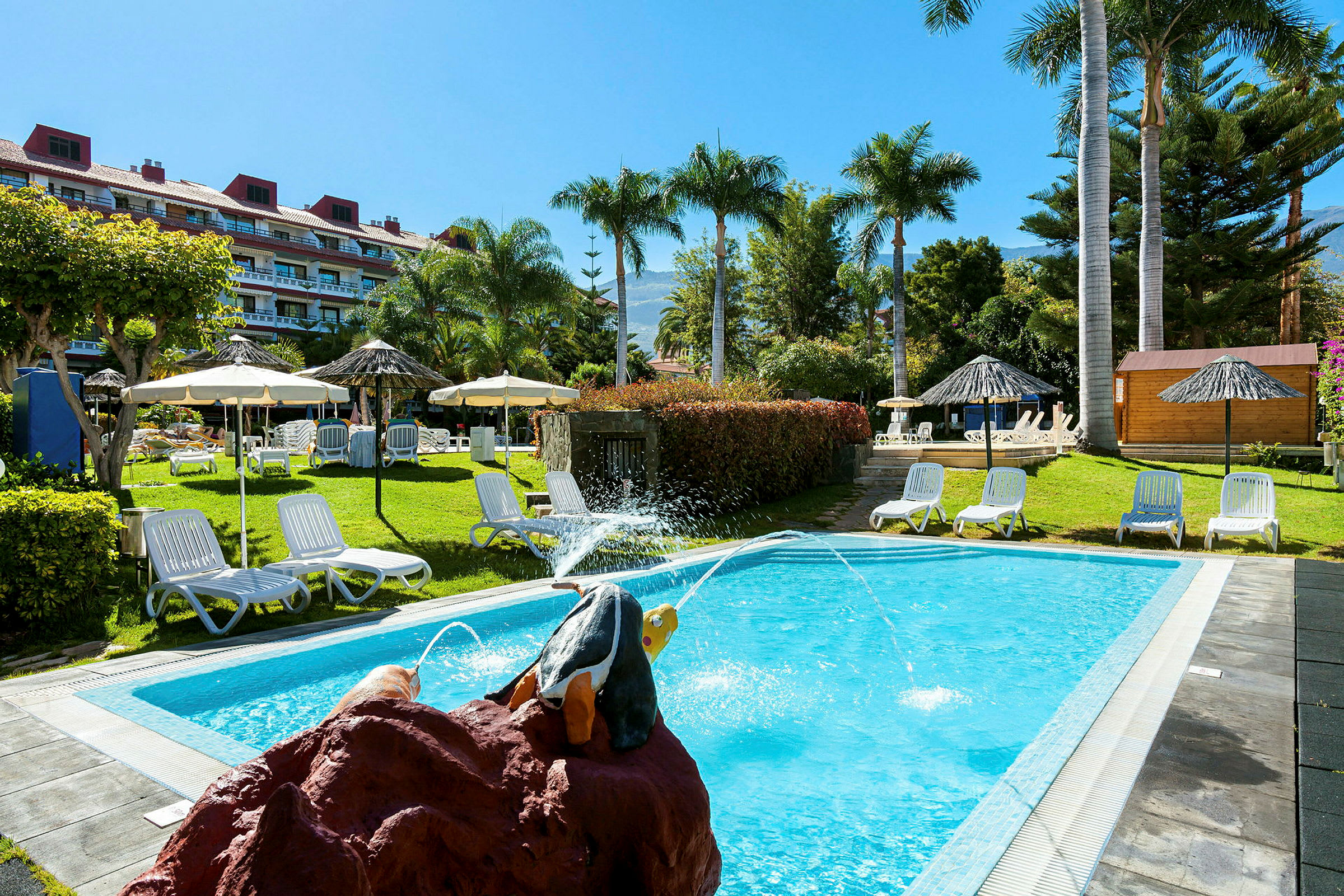 En pool med solstolar och parasoller på en grön gräsmatta. Bakgrunden visar ett hotell med flera våningar. (AI-genererad text)