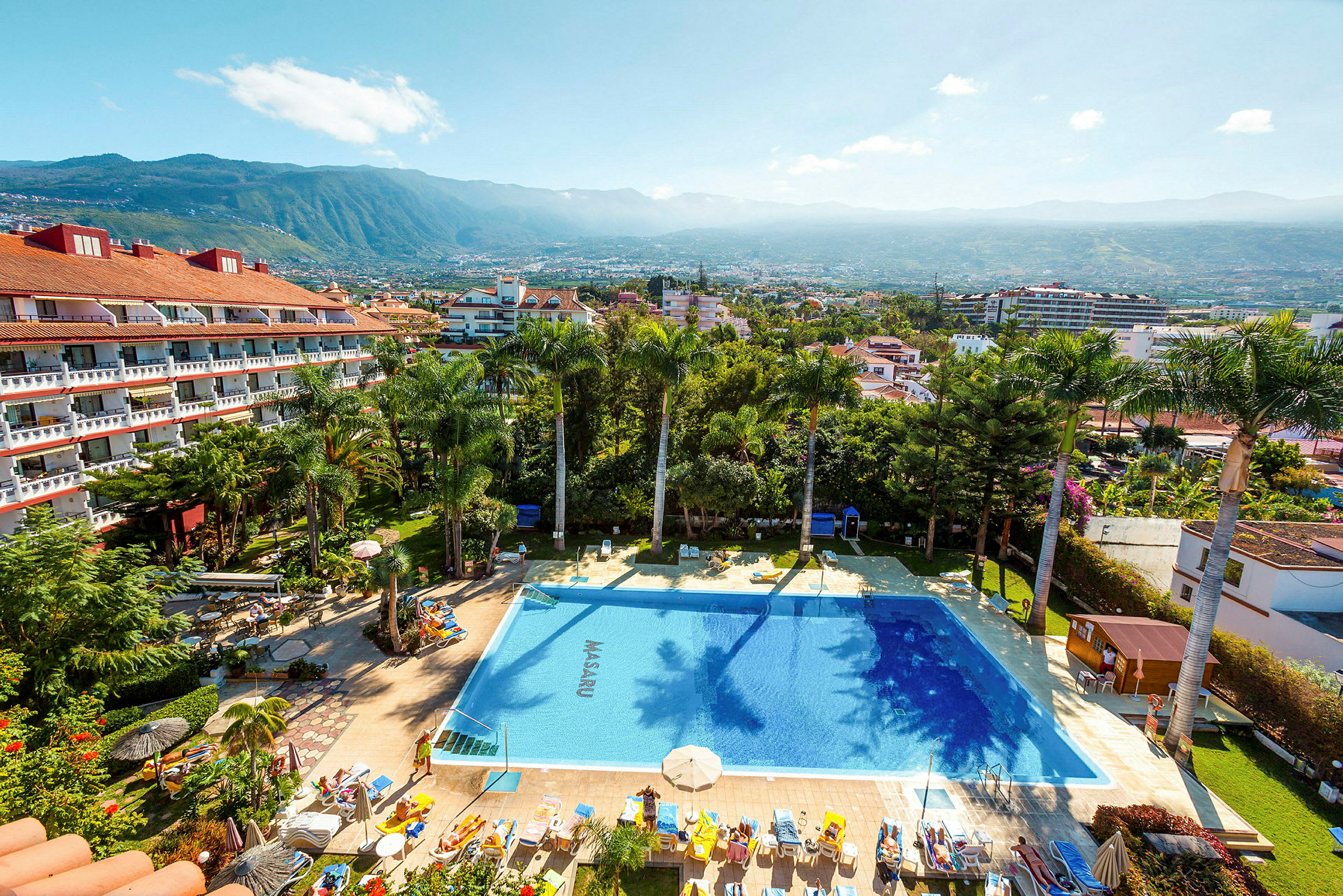 Vy över hotellområde med pool och omgivande trädgård, med utsikt över en stad och berg i bakgrunden. (AI-genererad text)