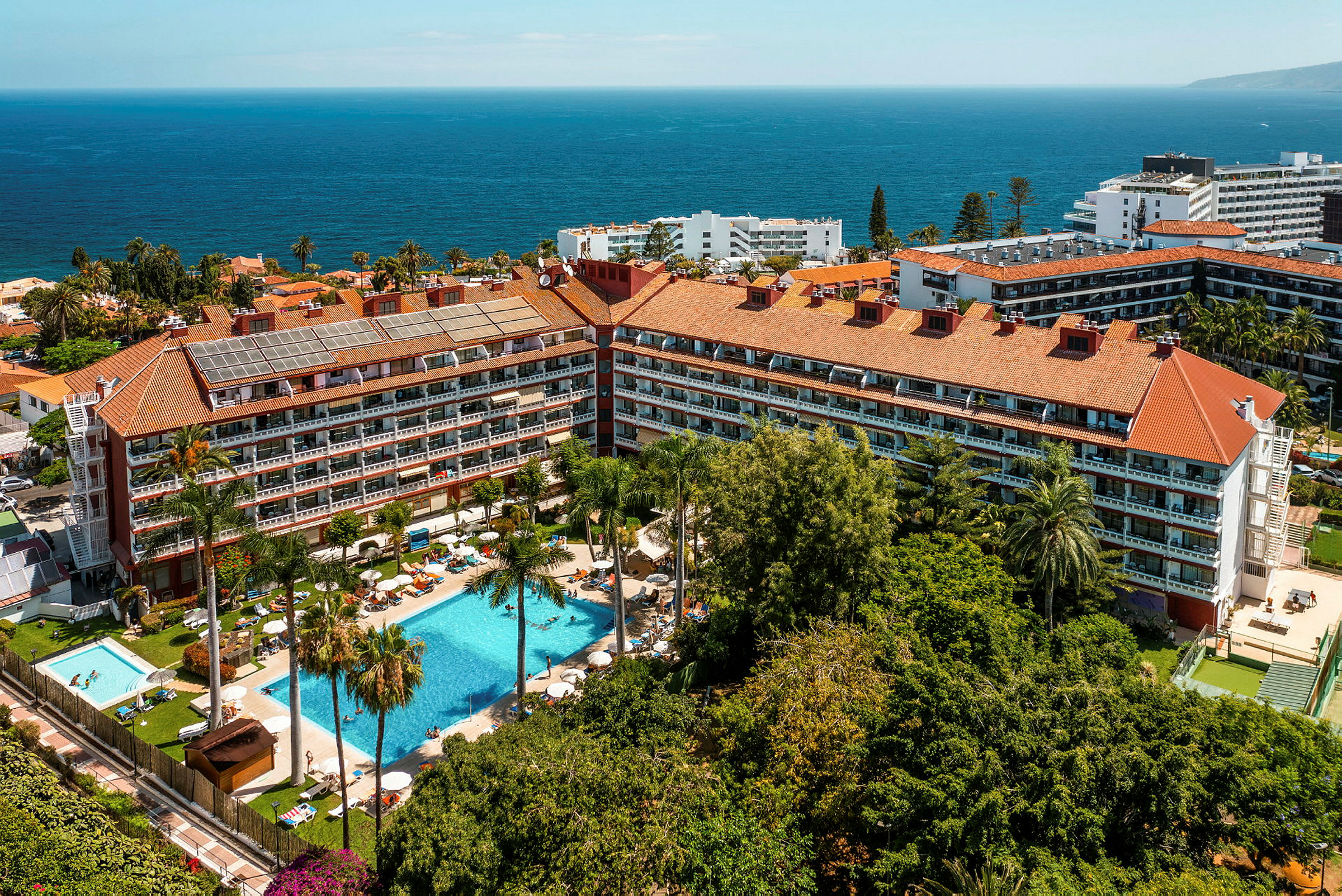 Flygfoto av ett hotellkomplex med pooler, palmer och havet i bakgrunden. (AI-genererad text)