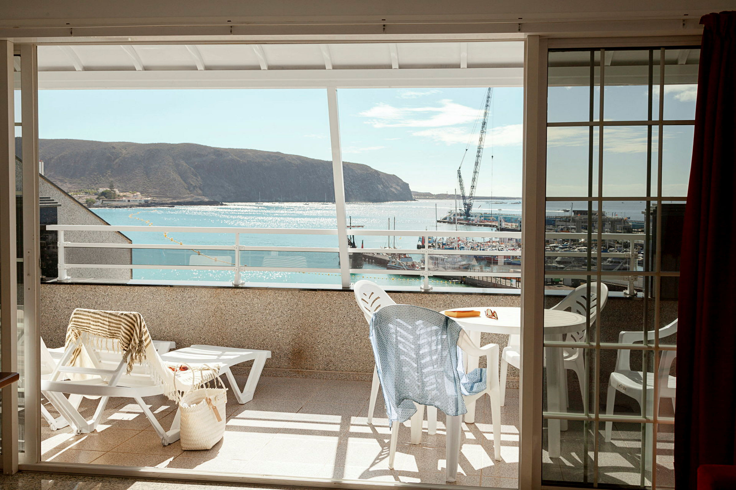 Utsikt från en balkong mot havet och en liten hamn, med utemöbler och solstolar på balkongen. (AI-genererad text)